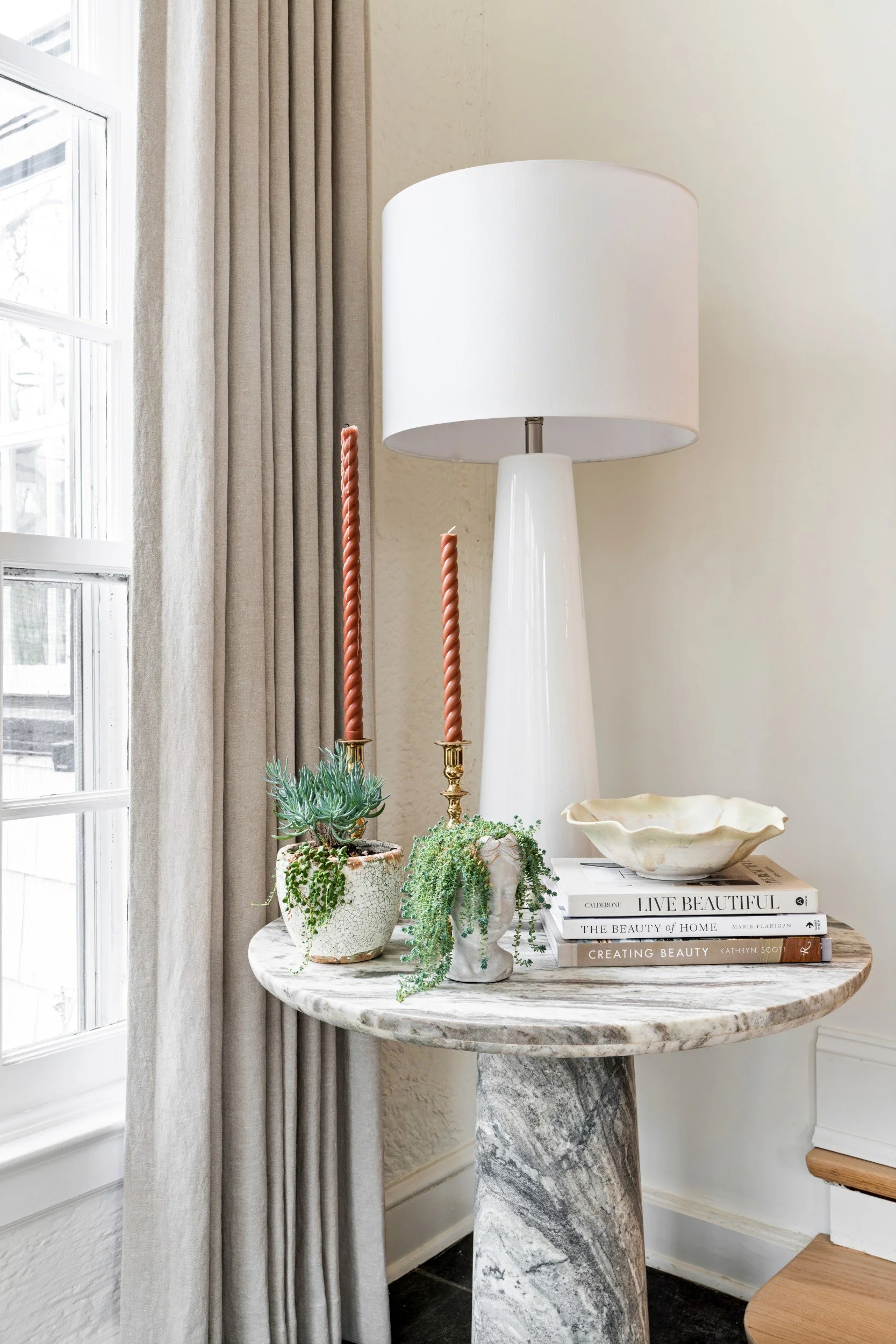 A round marble end table with small plants, a white glass lamp, rust colored twisted candle sticks and a stack of books.