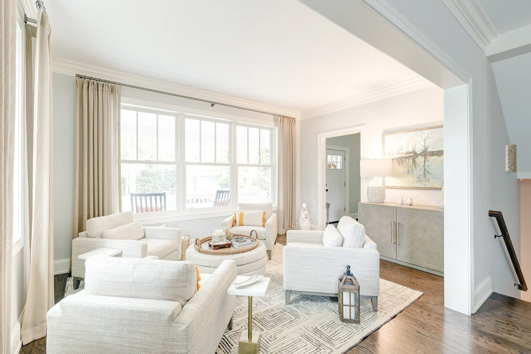 Bright living room with large windows, white furniture, a patterned rug, side table, and wall art.