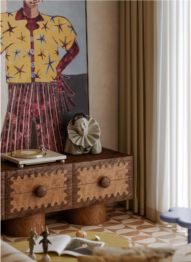Collected European interior featuring expressive artwork, an intricately carved wood chest, a vintage ceramic figure, layered curtains, and patterned tile flooring.