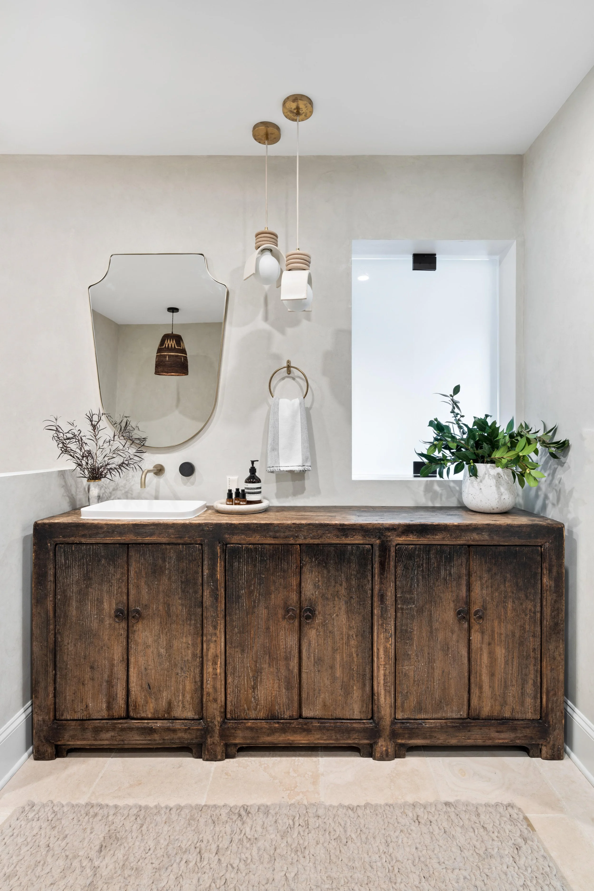 A bathroom vanity with a rustic wooden cabinet, a white sink, and decor including a vase with dried branches, a mirror, and small bottles. Above the vanity are haging light fixtures and a towel ring with a towel. There is a window with a plant in front of it and a beige rug on the floor.