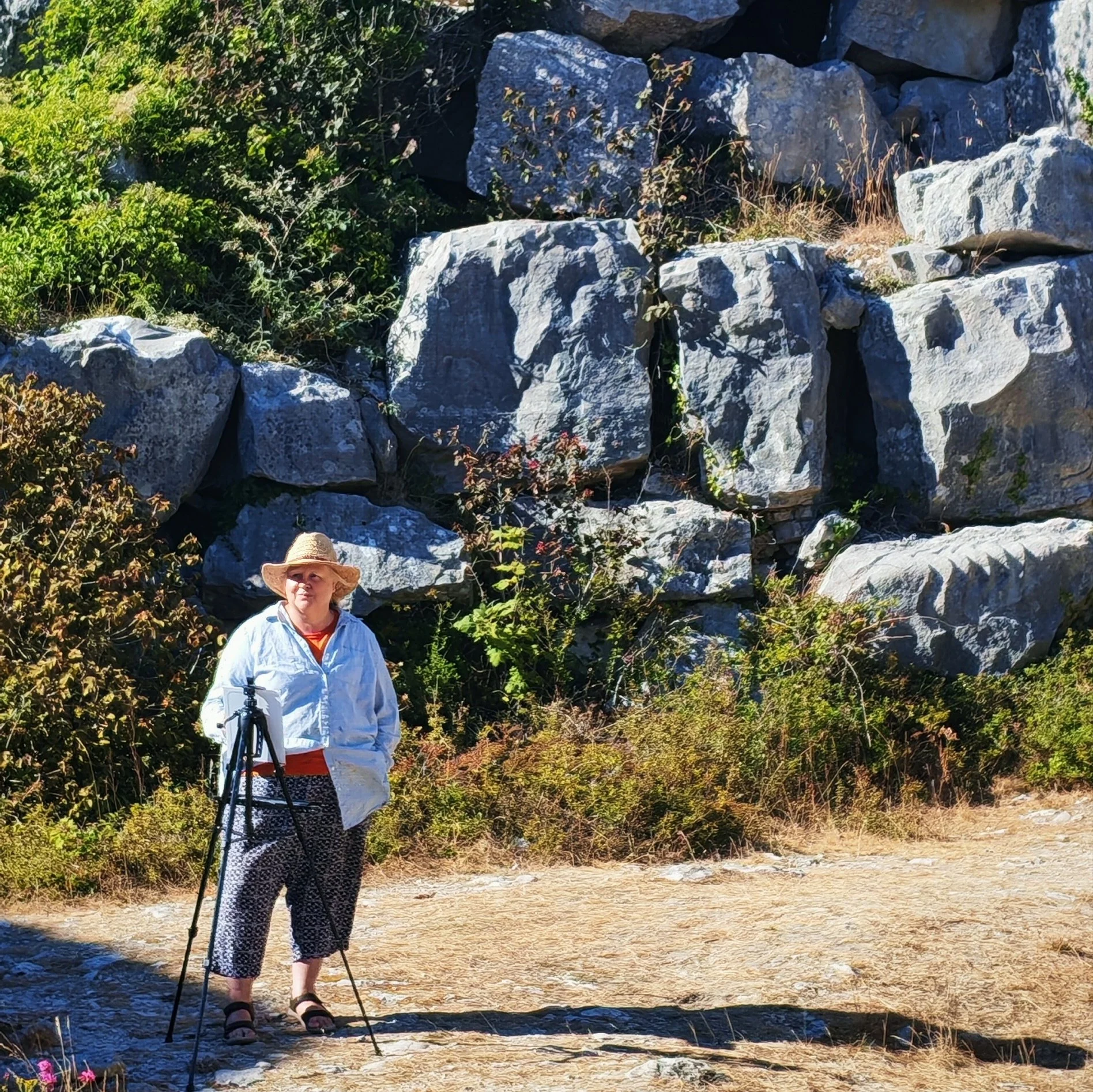 A woman wearing a straw hat, white blouse, and patterned pants standing outdoors with a painting easel, in front of a background of rocks, green foliage, and dry grass in a quarry setting.