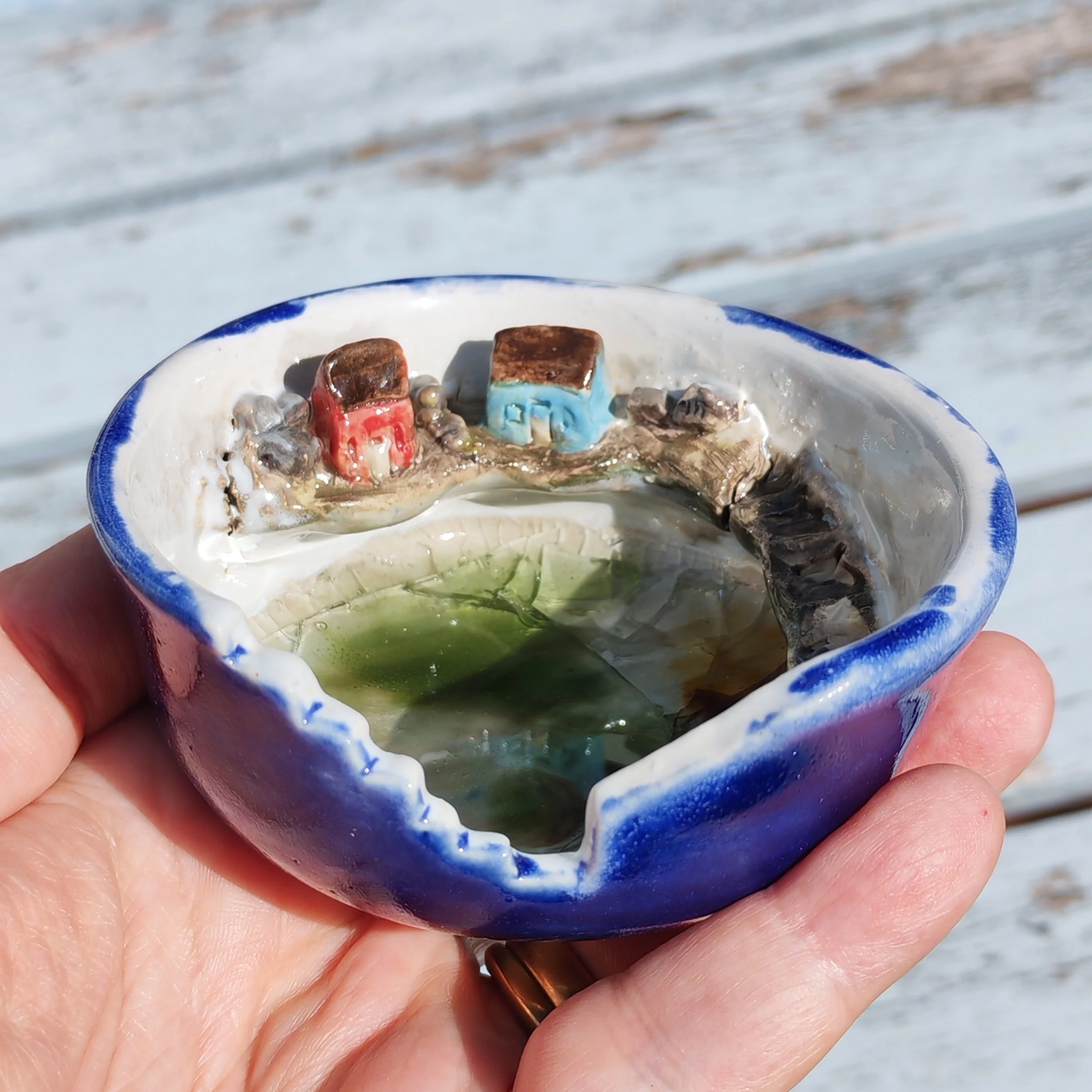 Person holding a small ceramic bowl containing ice cubes, several small colorful plastic toys, and water with ice, outdoors near a deck or boardwalk.