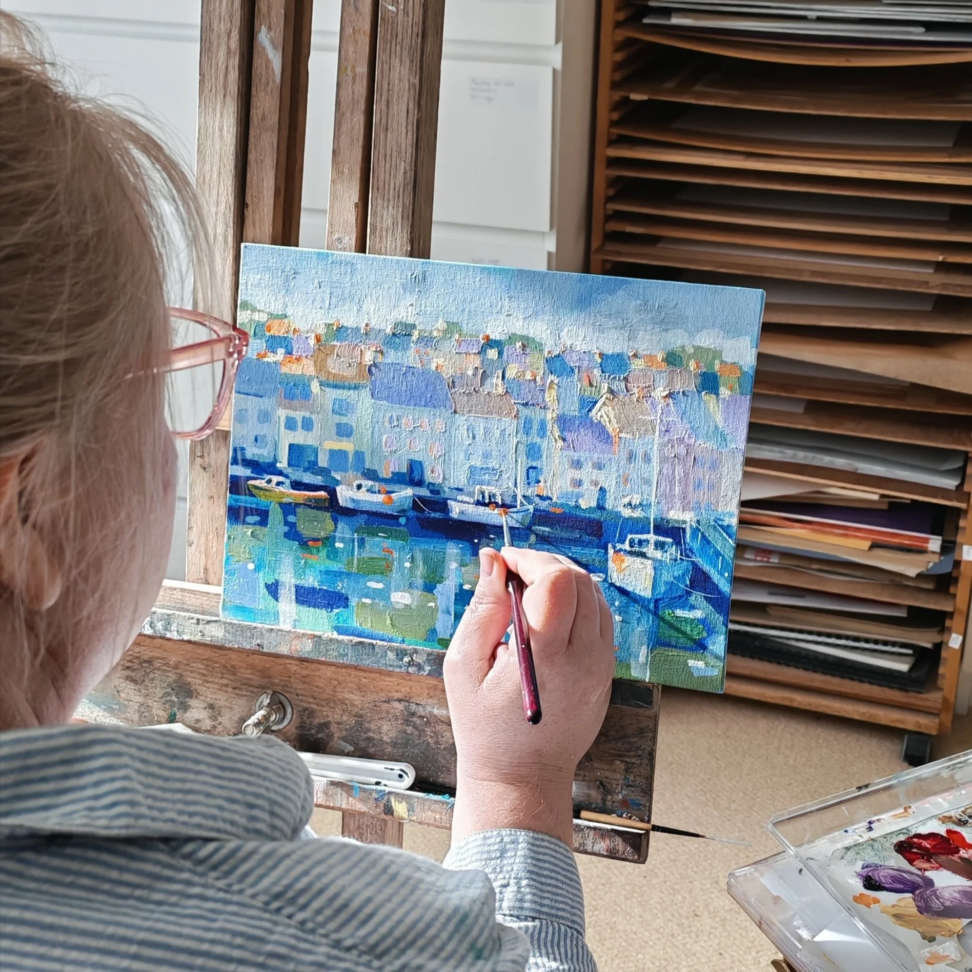 Person with glasses painting a colorful cityscape on a canvas, seated at a wooden table with art supplies, surrounded by shelves with stacked papers or canvases.