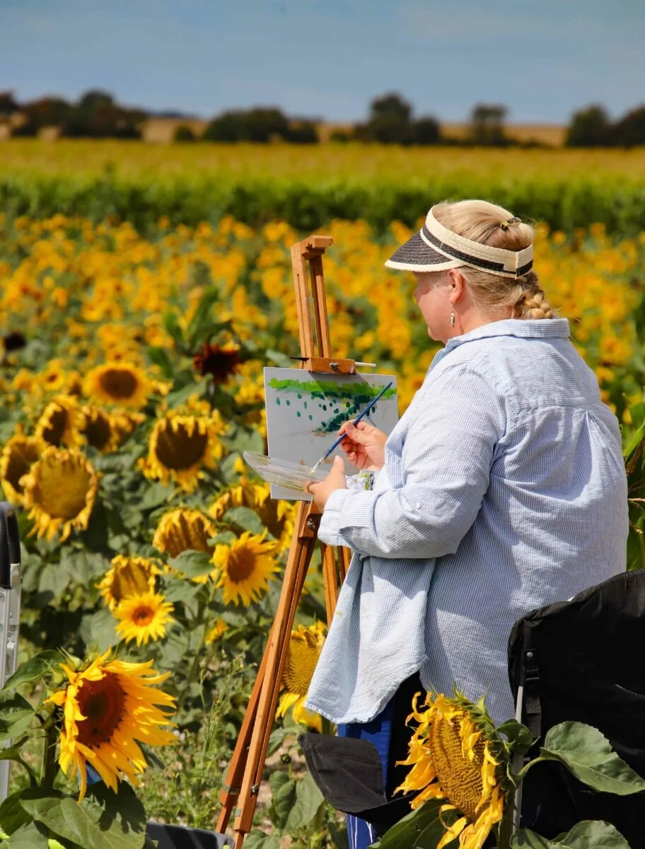 A woman painting a landscape of sunflowers on an easel in a sunflower field, wearing a wide-brimmed hat and light-colored shirt.