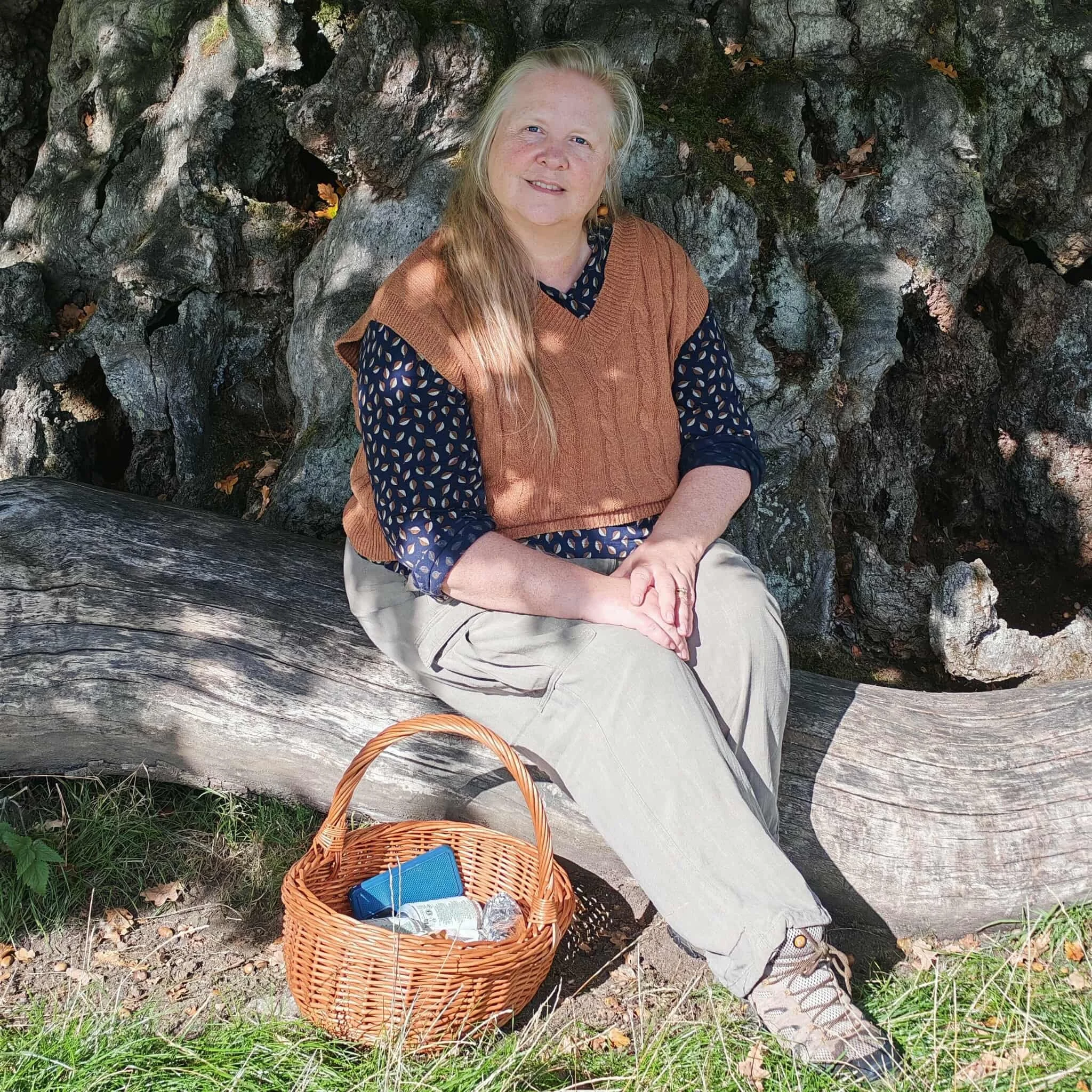 A woman with long blonde hair sitting outdoors on a rock formation, wearing a black blouse with a leaf pattern, a brown vest, and beige pants. A wicker basket with a blue book and a water bottle is placed on the ground nearby. The scene is surrounded by green grass and fallen leaves.