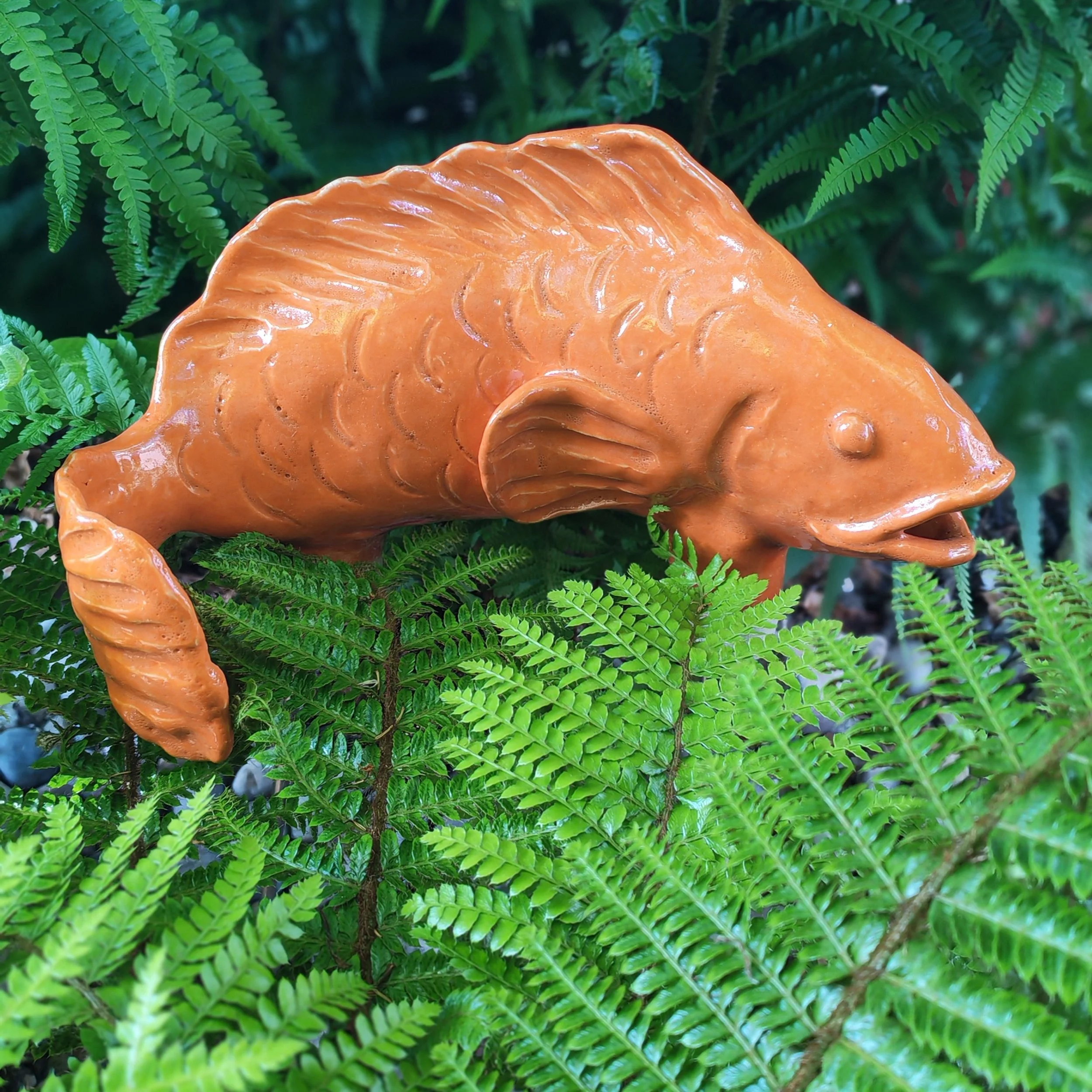 Close-up of a bright orange ceramic fish surrounded by green fern leaves.