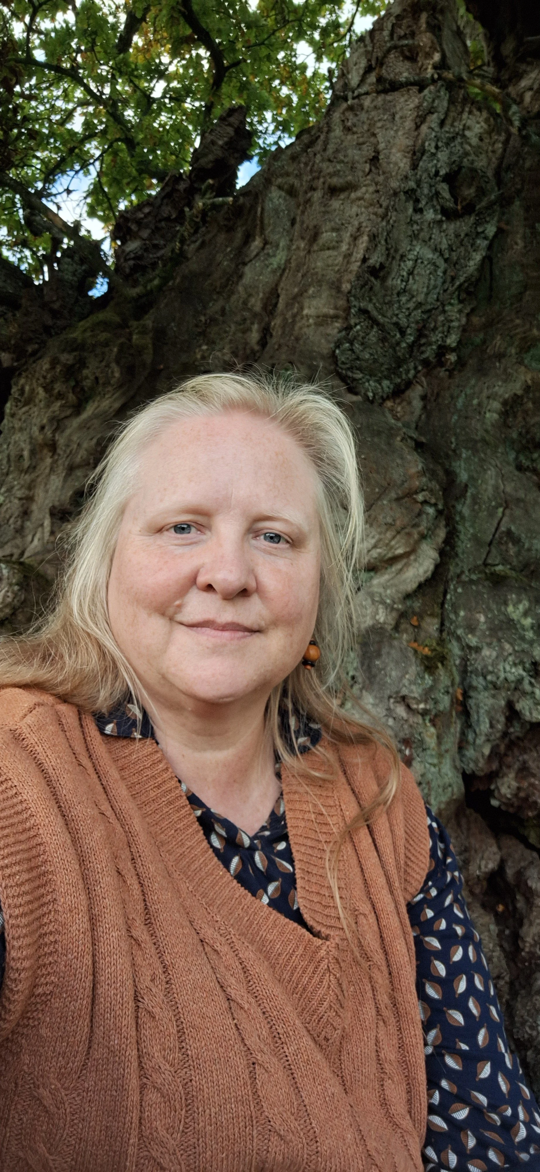 A woman with blonde hair and blue eyes standing in front of a tree with textured bark and green leaves, wearing a rust-colored sweater over a patterned blouse.
