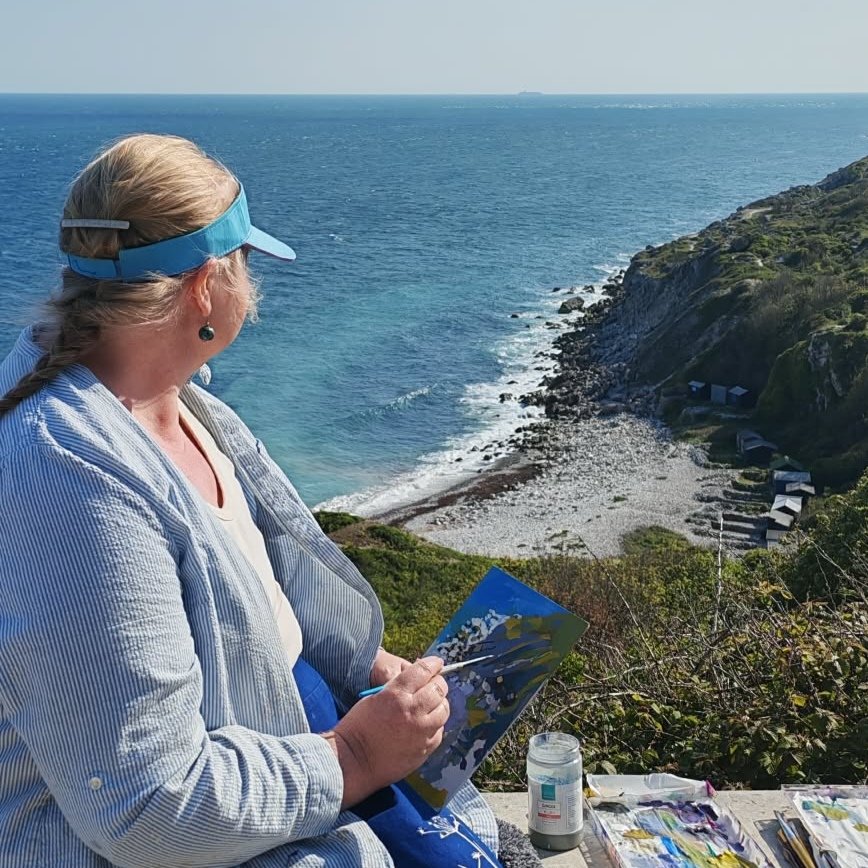 Woman with blonde hair, wearing a blue visor and striped shirt, painting outdoors on a cliff overlooking the ocean and rocky beach.