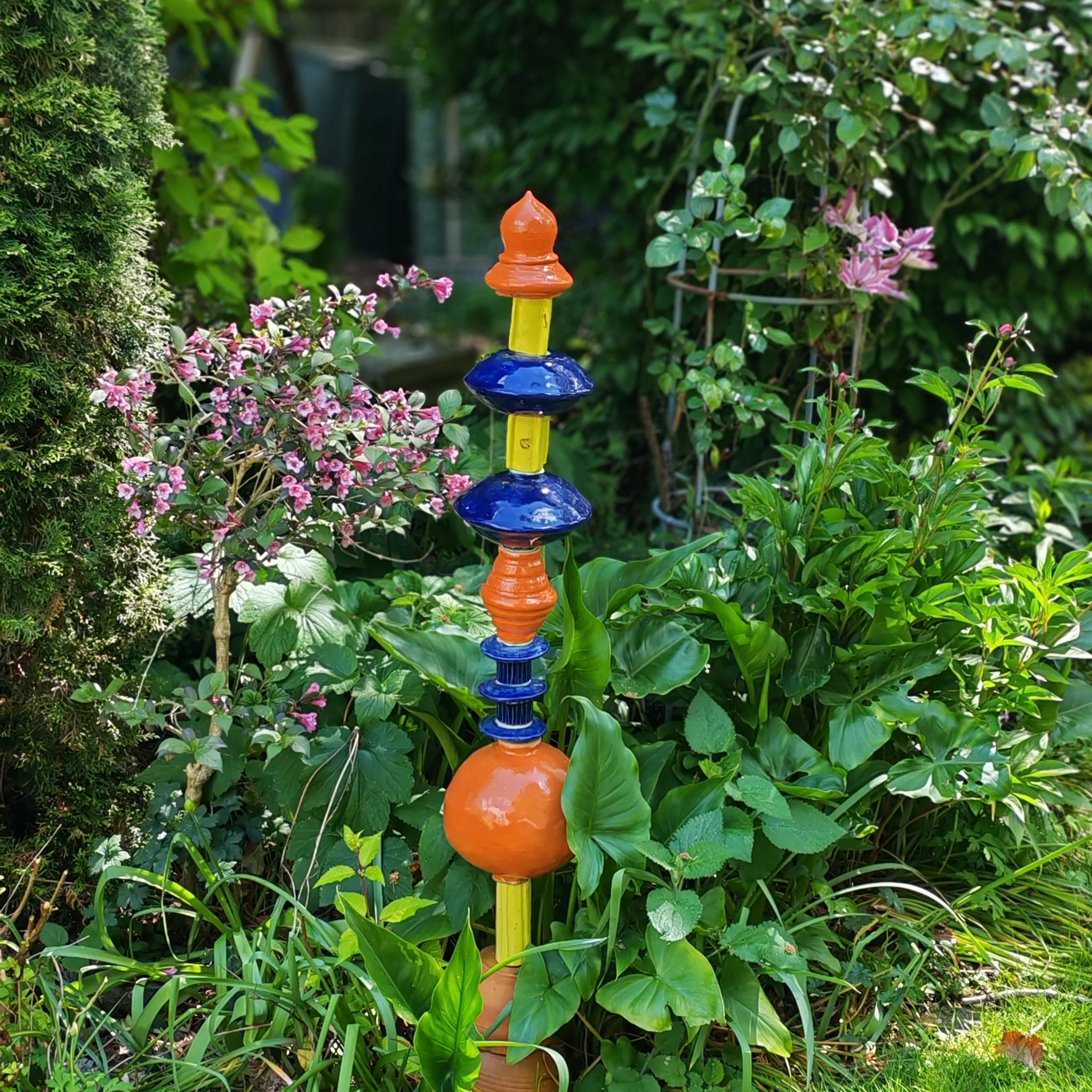 Colorful garden ornament with blue, orange, yellow, and green sections among green plants and pink flowers.