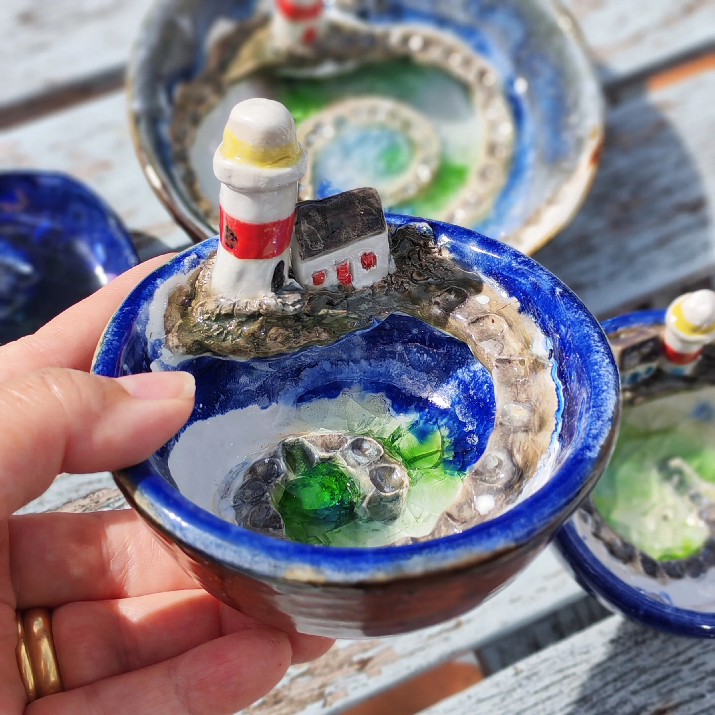 Close-up of a small decorative ceramic sailboat with a white sail with red stripes, held by a person's hand with a gold ring on the finger. The boat has blue and brown details and is set against a background of more ceramic boat decorations and a roc