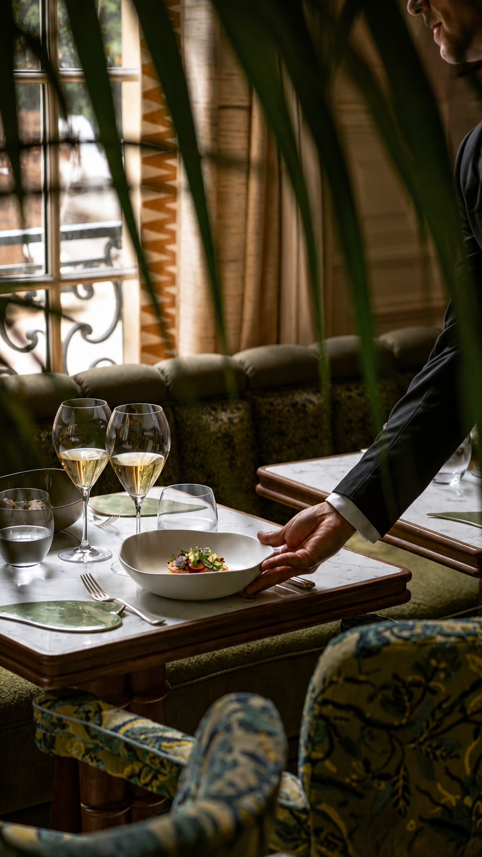 Un serveur dînant dans un restaurant élégant sert un plat dans une assiette blanche, tandis que des verres de vin blanc sont sur la table en marbre, avec un décor chaleureux et des rideaux en bois en arrière-plan.