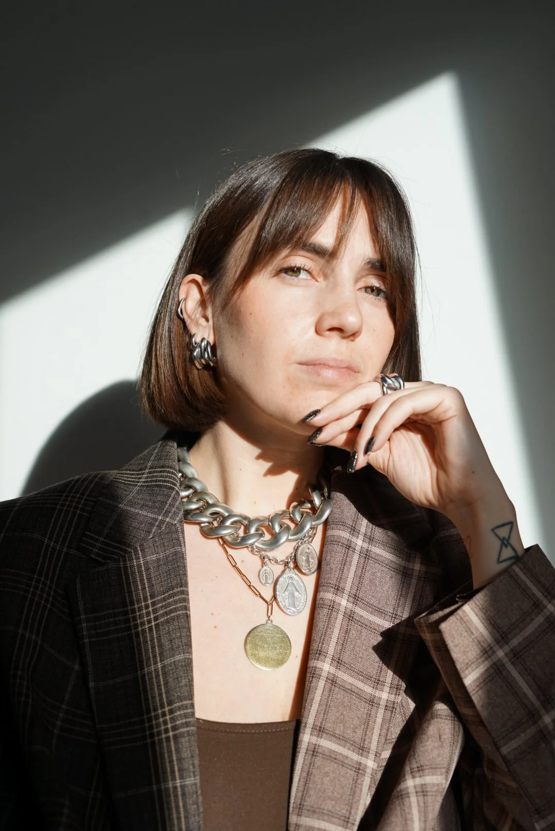 Une femme aux cheveux bruns courts portant des bijoux en métal, dont un collier épais, plusieurs pendentifs et boucles d'oreilles, vêtue d'une veste à carreaux, sous un éclairage laissant une ombre géométrique sur le mur blanc derrière elle.
