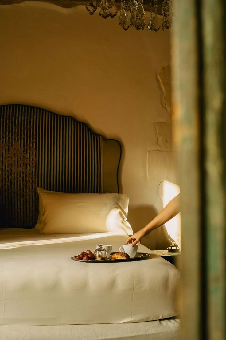 Un lit avec un tête de lit en bois stylisée, oreillers, plateau avec petit-déjeuner comprenant des croissants, des fruits et des tasses, dans une chambre chaleureuse avec lumière douce.