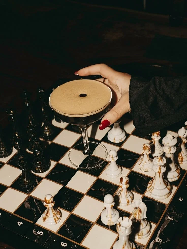 Une main tenant un cocktail noir dans un verre à martini, sur un échiquier avec des pièces d'échecs.