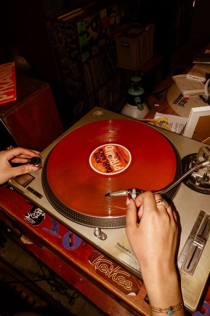 Une personne manipule un tourne-disque avec un disque vinyle orange. La main droite tient la tête de lecture, tandis que la main gauche ajuste un bouton. Le tourne-disque est posé sur une table en bois avec des autocollants, dans un environnement qui semble être un magasin de musique ou une salle de DJ.