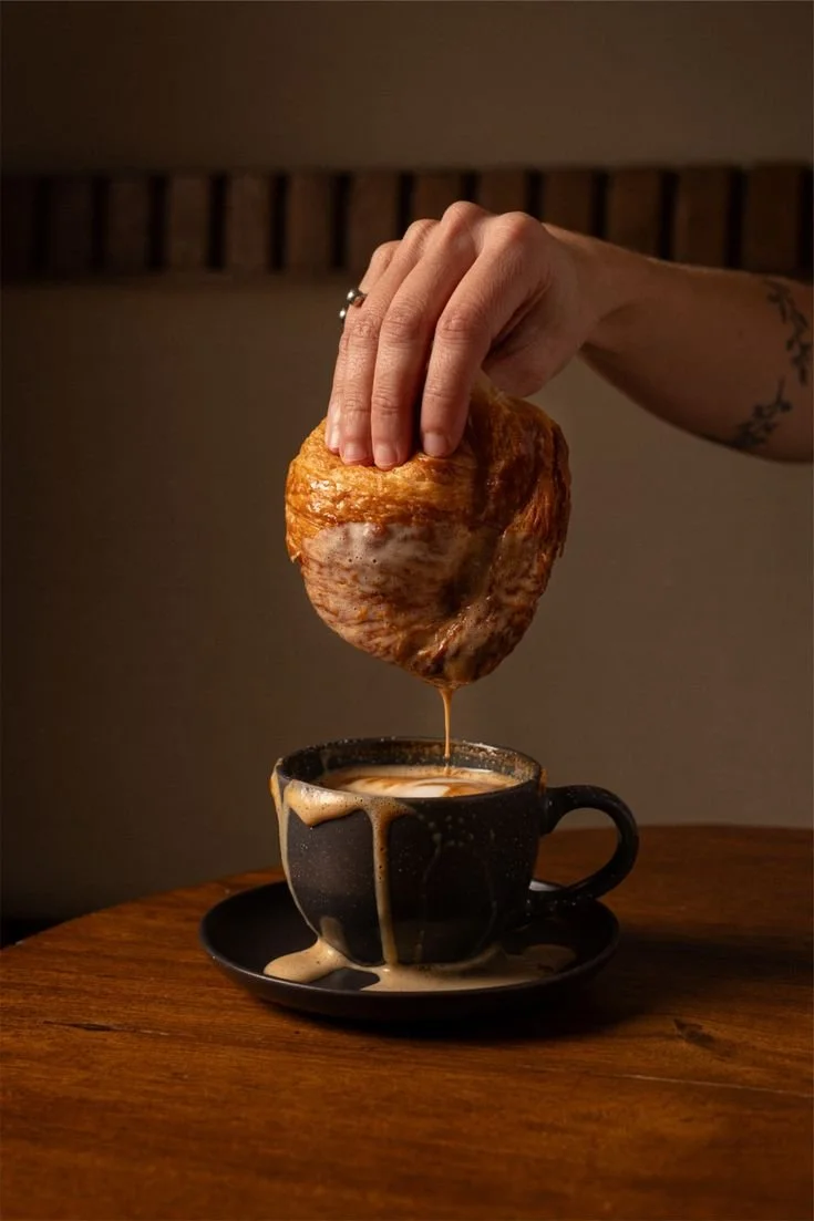 Une main tient un morceau de poulet frit au-dessus d'une tasse de café, avec de la sauce qui coule sur la tasse et la table en bois.
