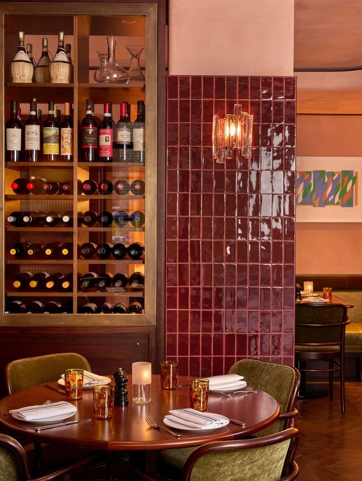 Une salle à manger dans un restaurant avec une table ronde, des chaises en velours vert, des verres, des assiettes, des couverts, une bougie et une bouteille de sel. En arrière-plan, un mur en carreaux rouges avec un éclairage décoratif et un mur en bois avec une étagère à vin.