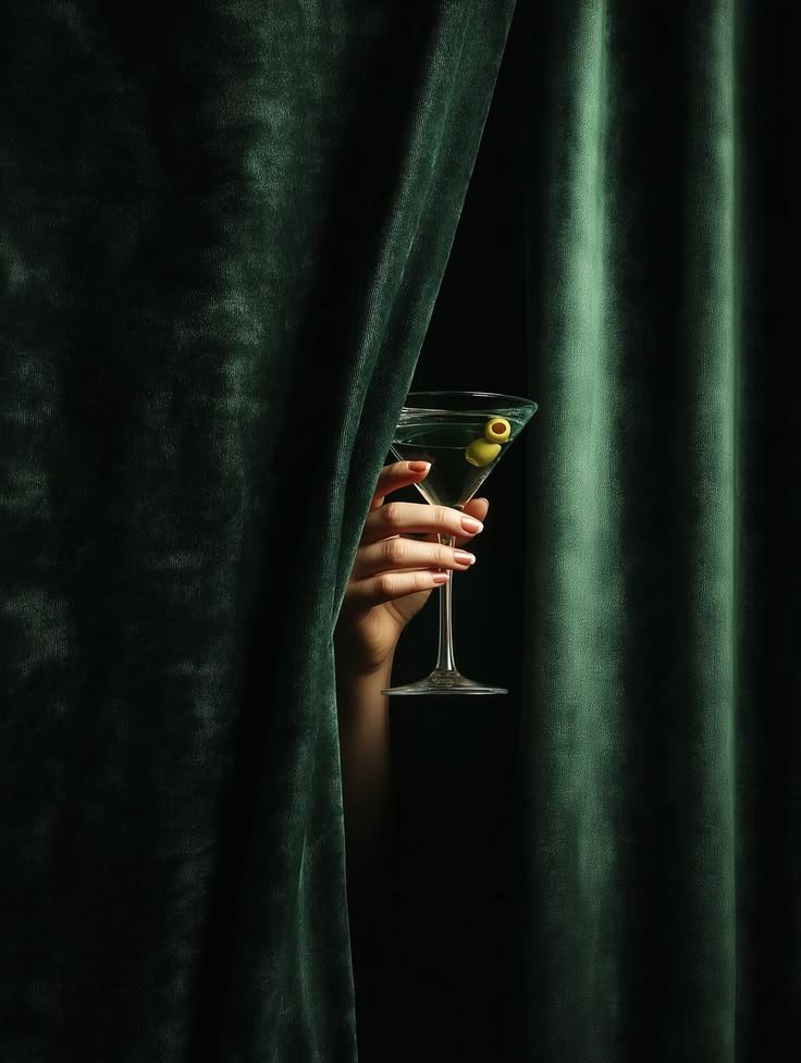 Une main tient un verre à cocktail derrière un rideau vert. Le verre contient des olives vertes