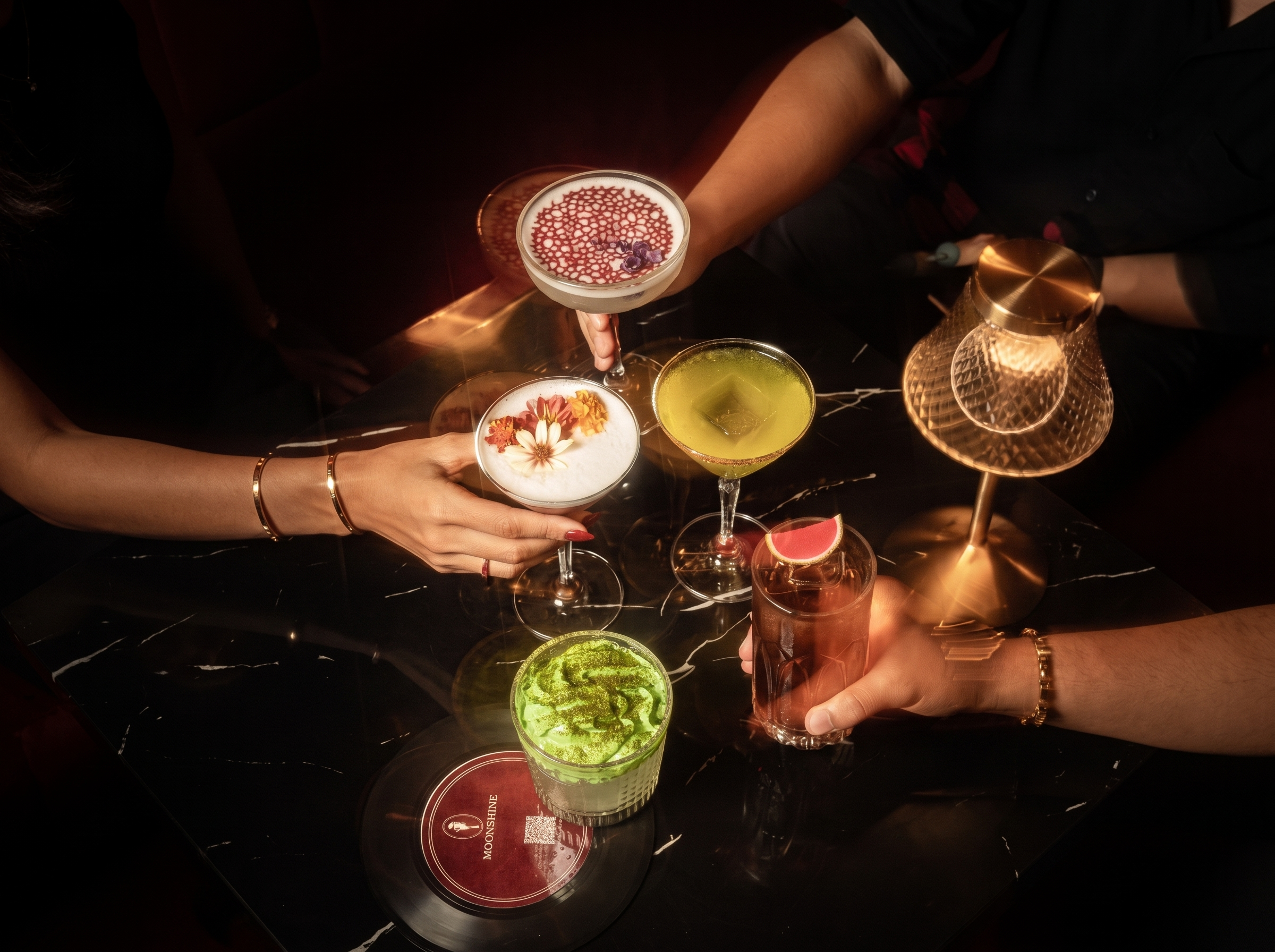 Plusieurs cocktails colorés avec des décorations florales, dans un environnement sombre, avec une personne portant des bracelets en or.