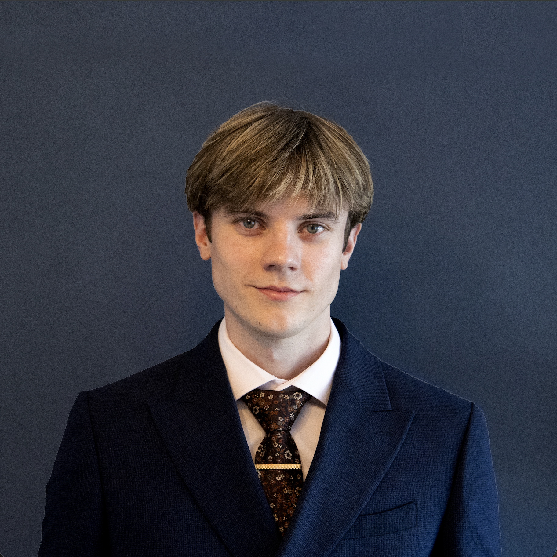 A young man in a dark suit, white shirt, and patterned tie posing against a dark background.