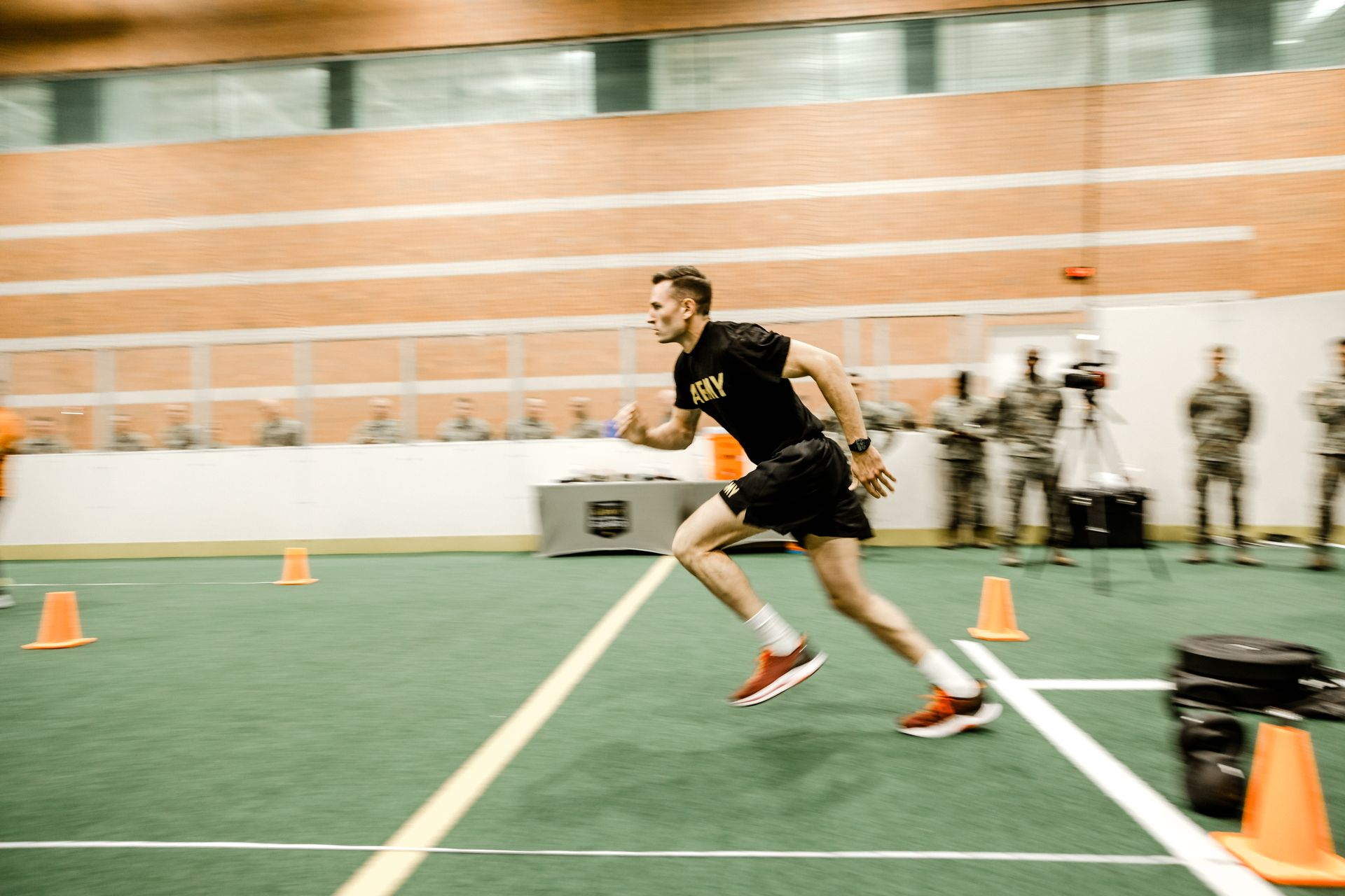 A man in a black shirt and shorts running indoors, wearing a watch, with orange cones set up for a training course, and military personnel in the background.