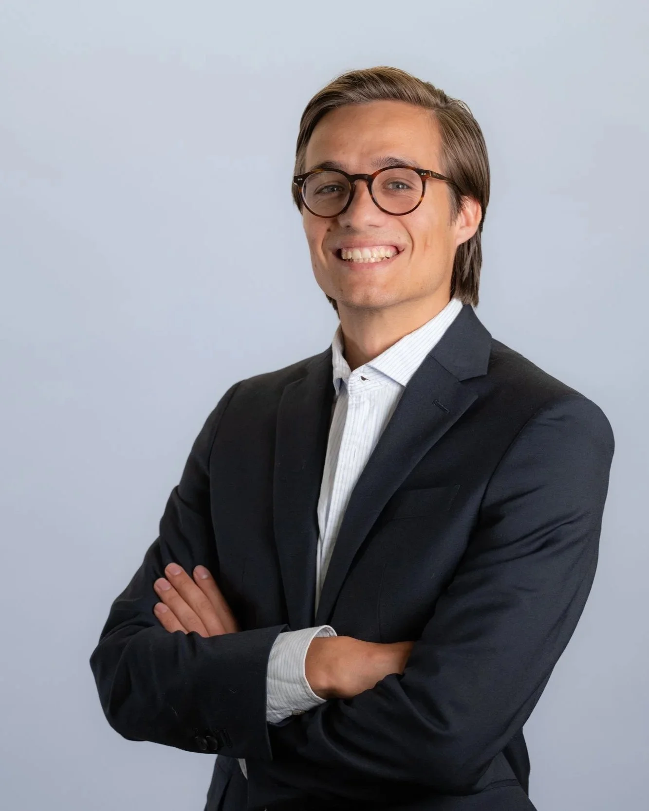 Portrait of a young man with glasses, wearing a dark suit and white shirt, smiling, posing with arms crossed against a plain light blue background.