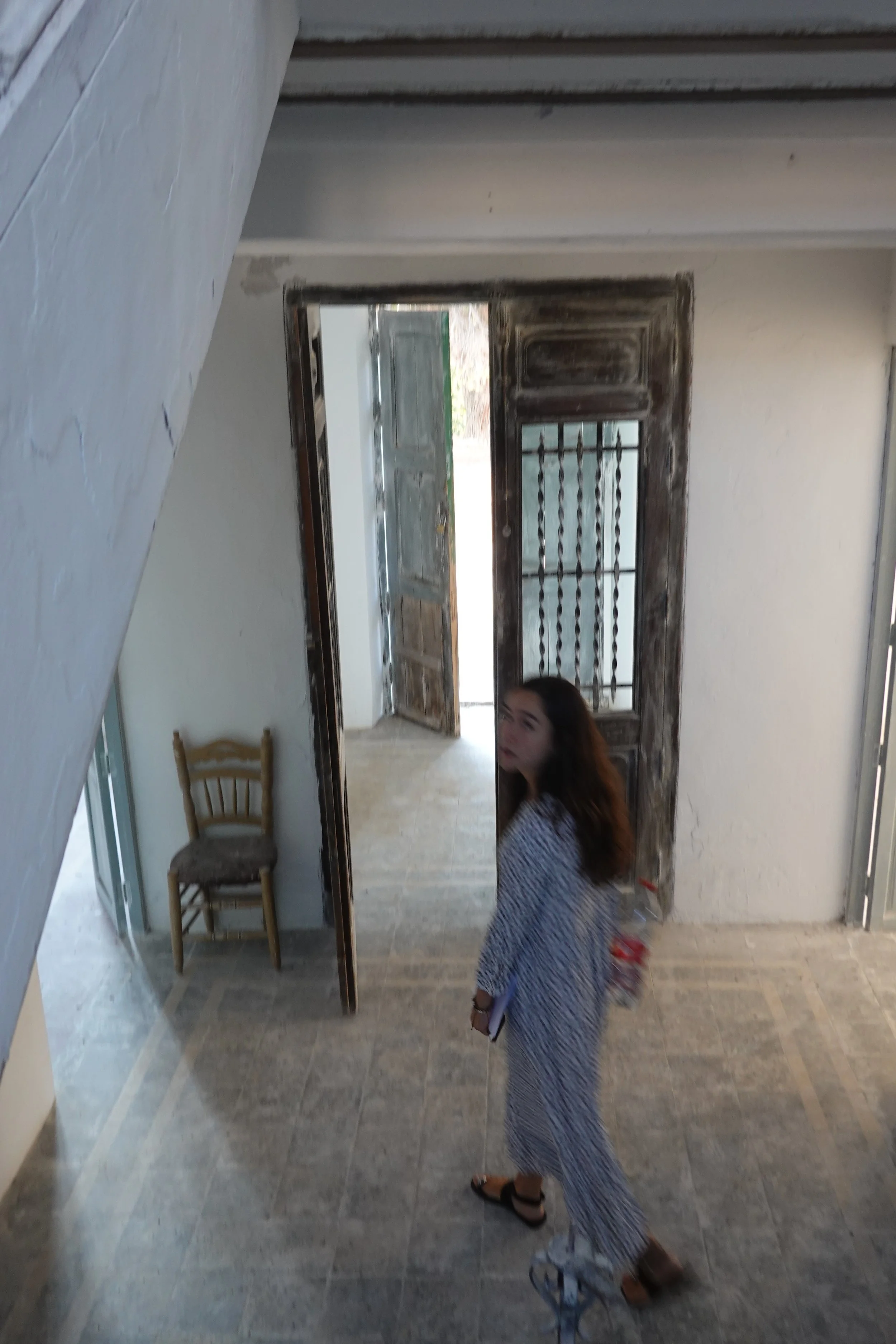 Mirror reflecting a woman with long hair wearing a blue and white patterned dress, standing indoors near a wooden chair and an open door showing an outside area.