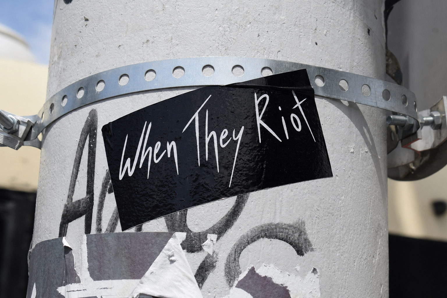 A black sticker with white handwritten-style text that reads 'When They Riot' is attached to a white utility pole with a metal band and other torn stickers.
