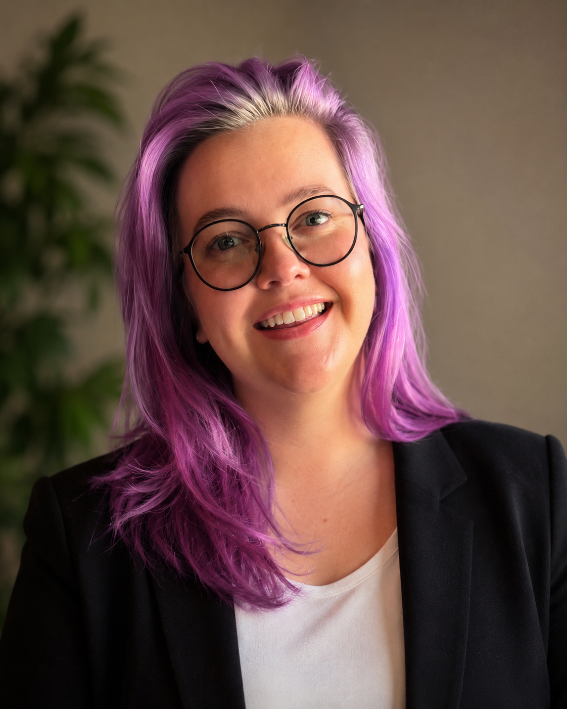 Smiling woman with purple hair and glasses, wearing a black blazer, indoors with a blurred plant background.