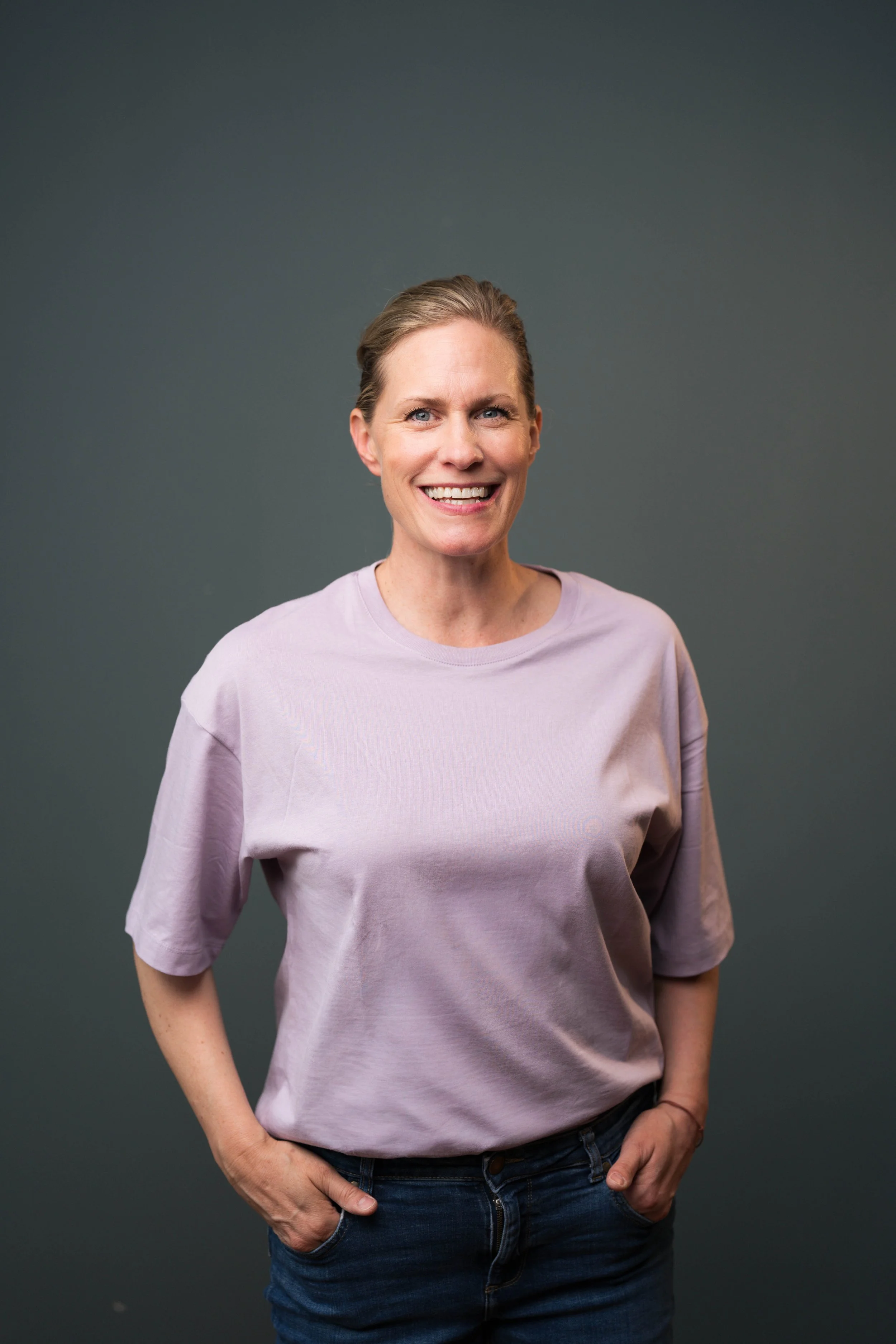 Porträt einer lachenden Frau mit kurz braunen Haaren, grauem Hintergrund, lässiges rosa T-Shirt und Jeans, die eine Hand in die Tasche steckt.