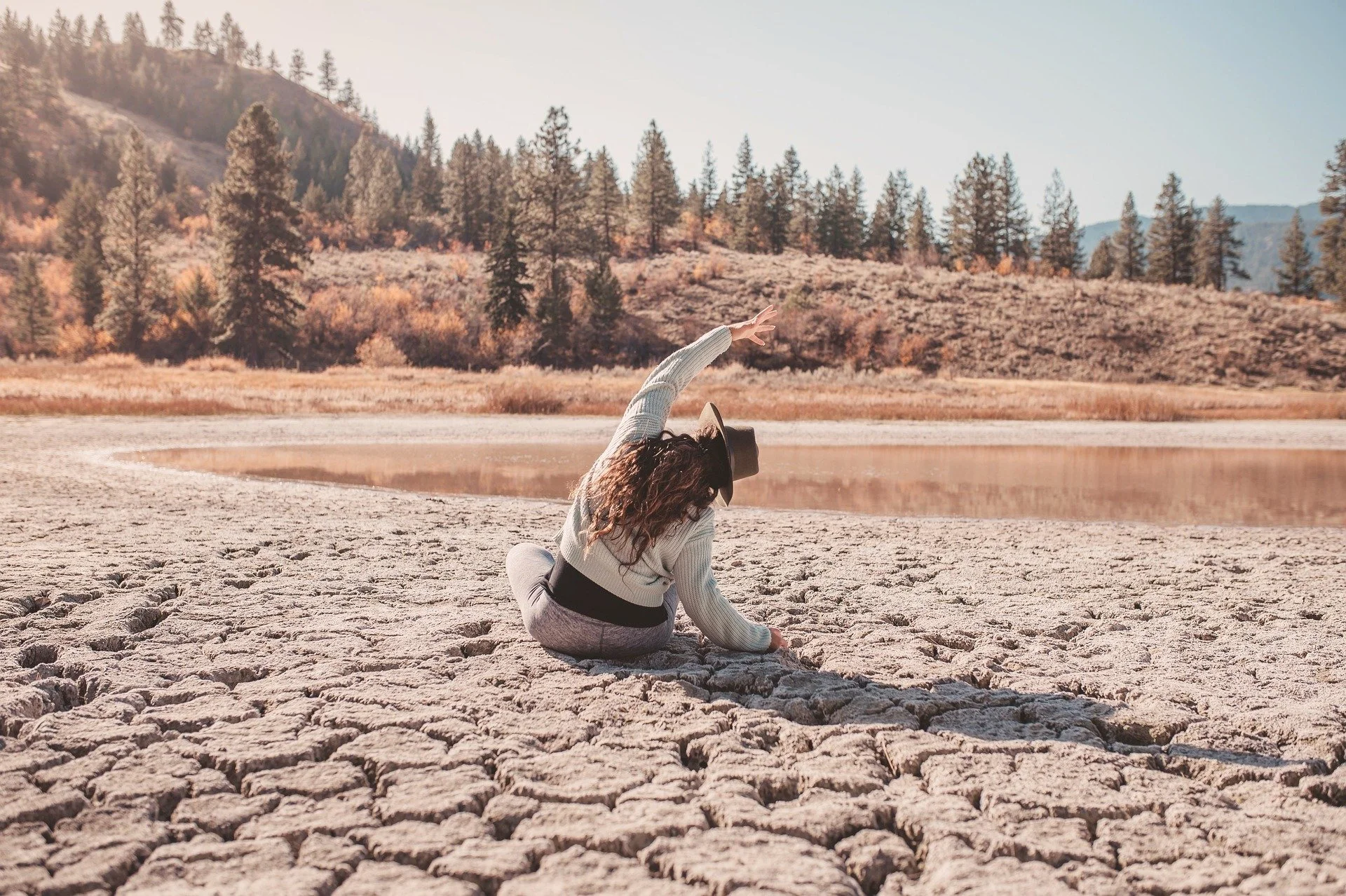 A yoga student with curly hair, wearing a hat, gray pants, and a white sweater, is sitting on cracked dry ground near a body of water, reaching out with her right arm, in a natural outdoor setting with trees and hills in the background during sunset.