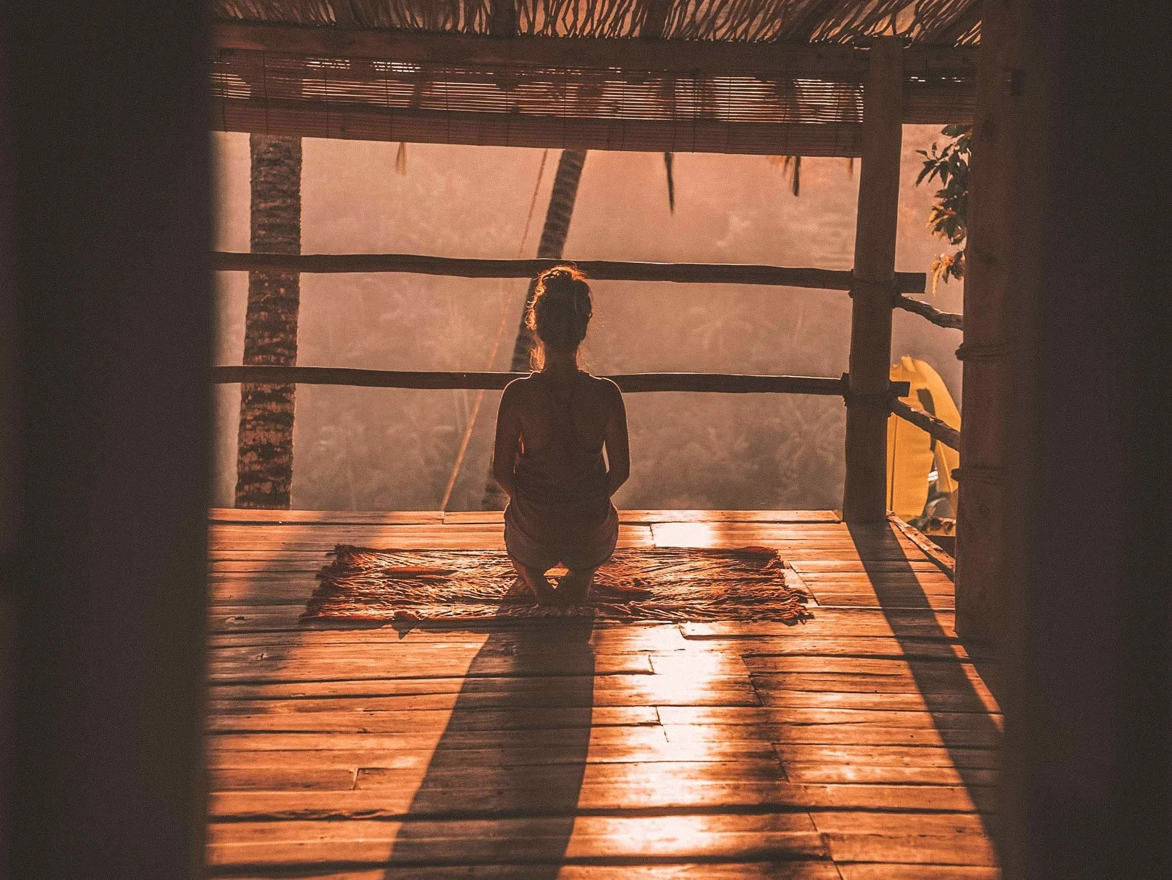 A yoga teacher sitting on the wooden floor of a yoga shala at sunset, facing away and looking towards the horizon where the sun is setting over water. The silhouette is framed by a rustic wooden structure and tropical plants.