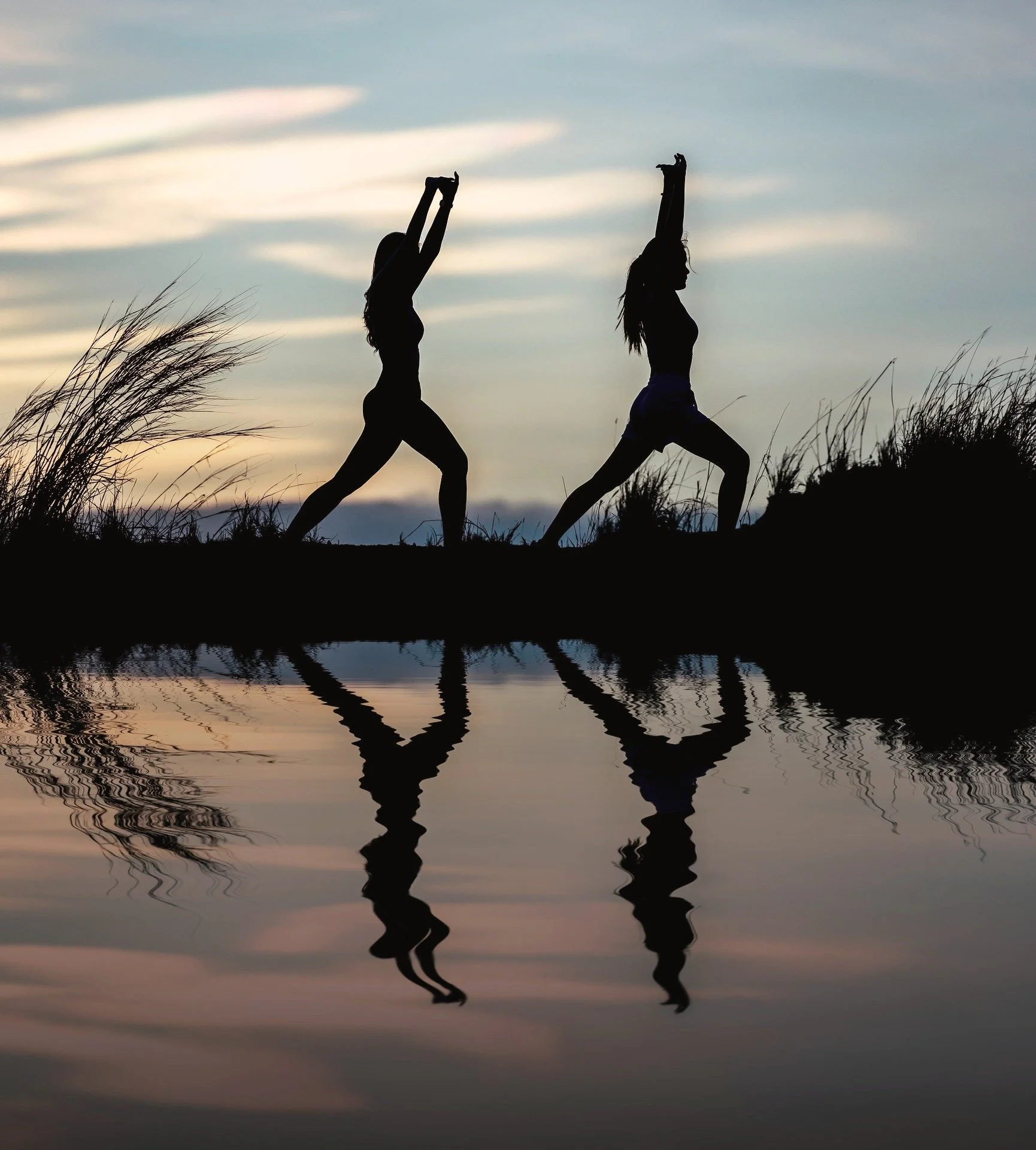 Silhouettes of two women stretching with arms raised, standing near water at sunset, with their reflections visible in the calm water.