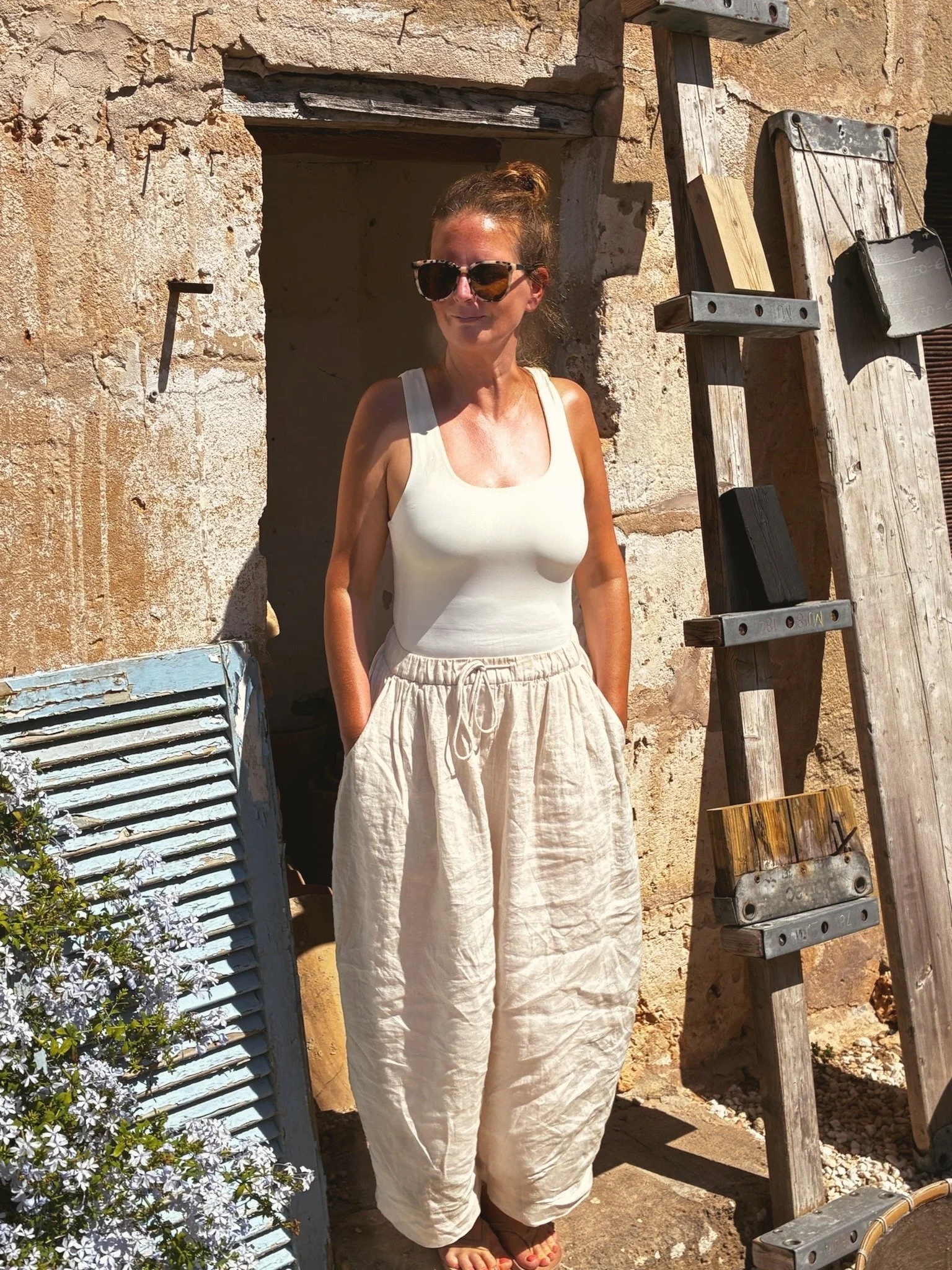 A woman wearing sunglasses, a white tank top, and loose white pants standing outdoors against an old textured wall with wooden and metal objects nearby.