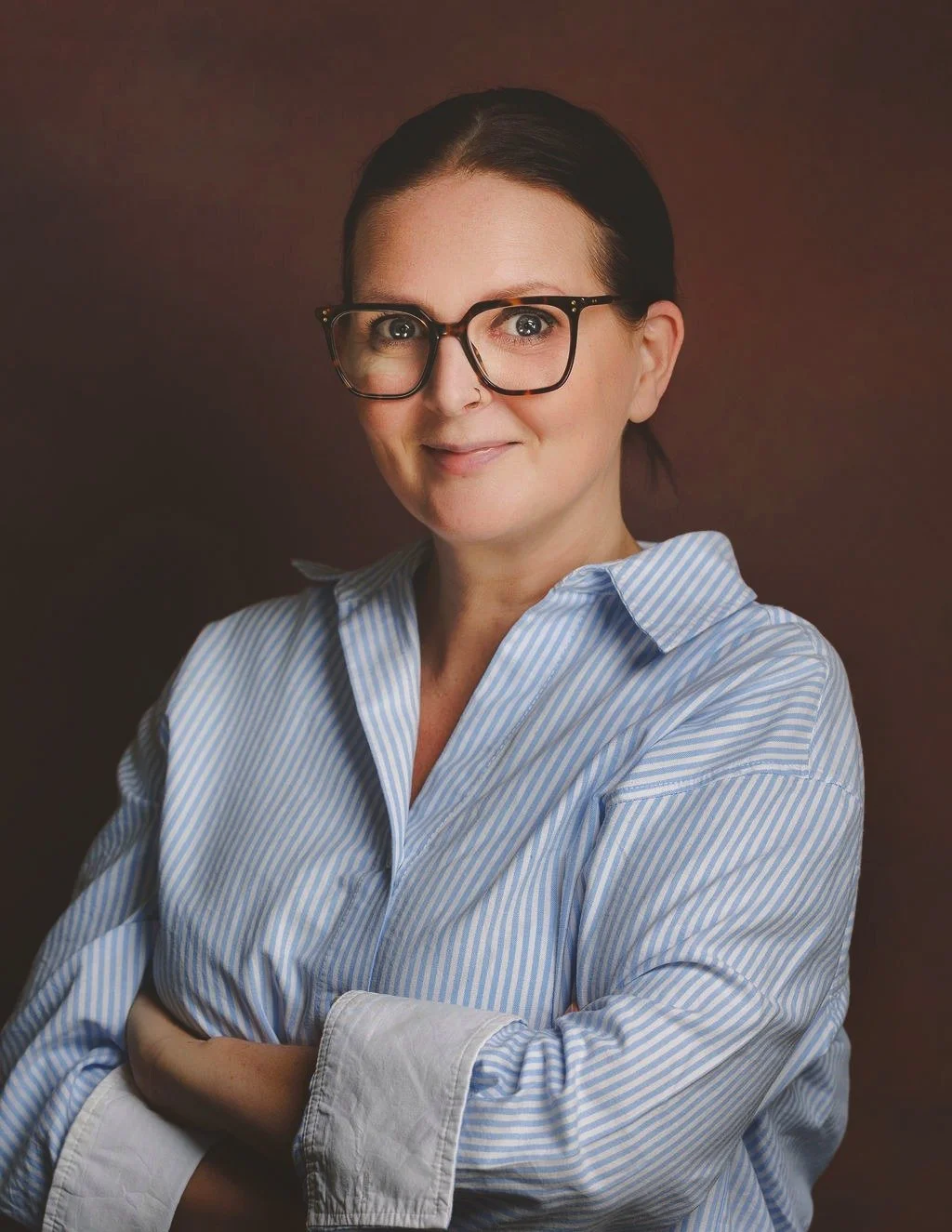 A woman, Sara Dickenson, with dark hair, wearing glasses and a blue and white striped shirt, smiling against a brown background.