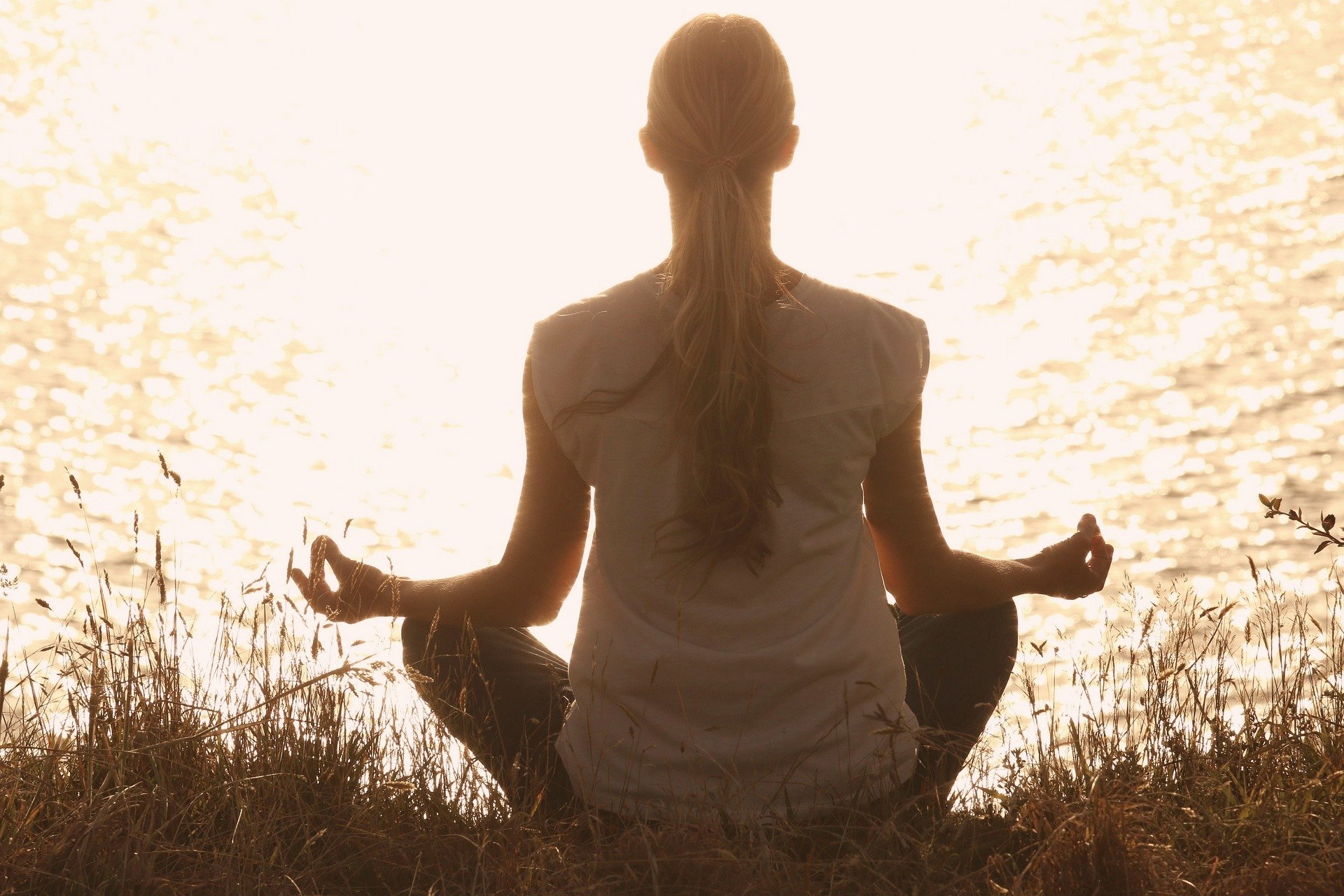 A person practicing yoga in a seated meditation pose by the water at sunset, facing away from the camera, with long hair tied back.