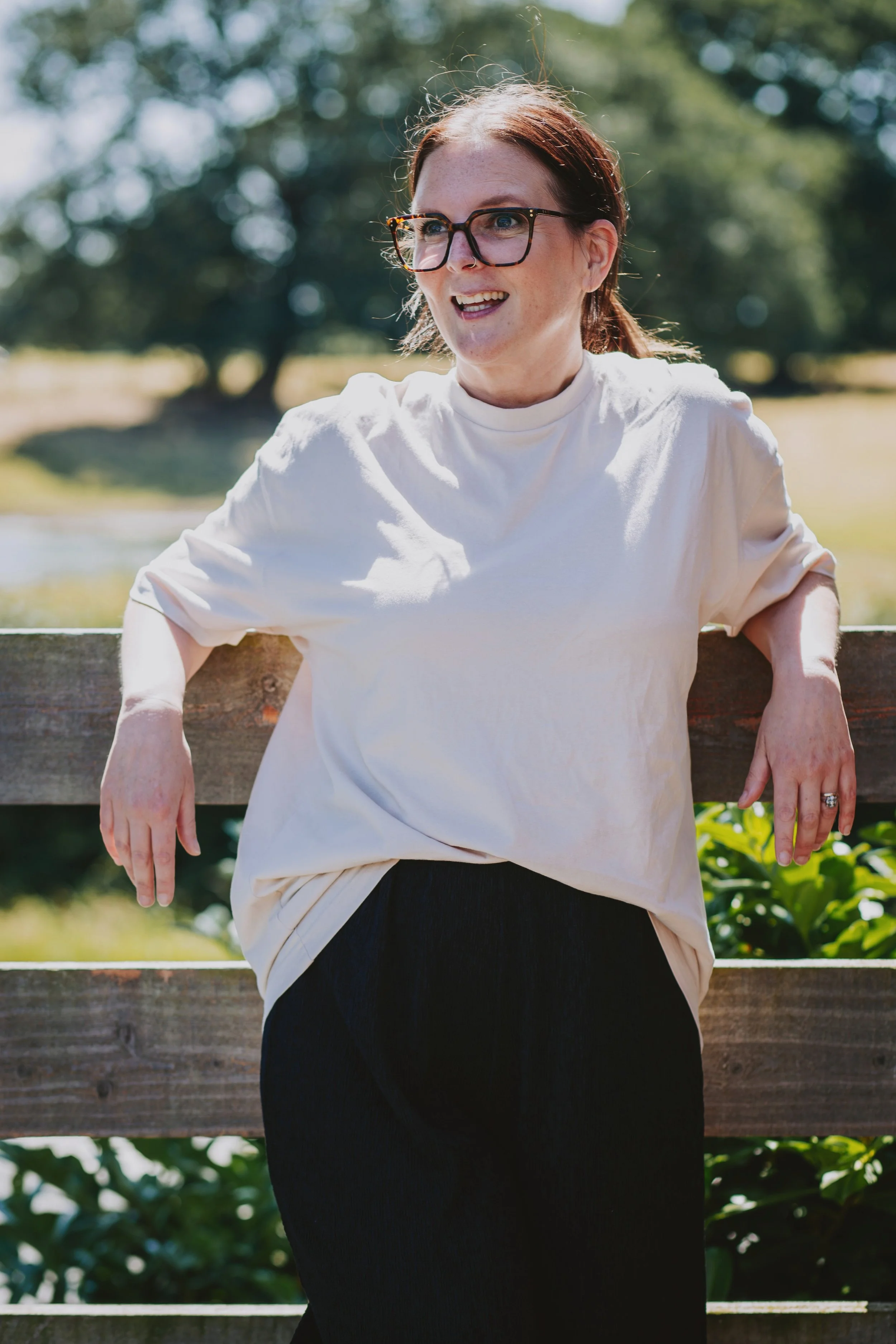 A woman with reddish-brown hair, glasses, and a white t-shirt sitting outdoors on a park bench, smiling and leaning slightly to the right.