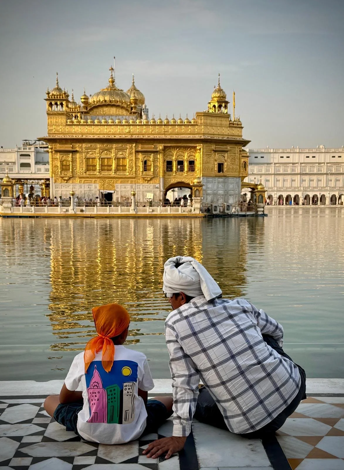 A man and a child sitting by the water, looking at the Golden Temple in Amritsar, India.