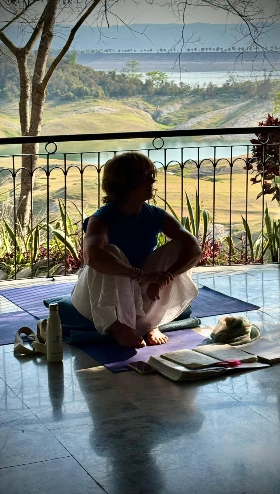 A yoga teacher sitting on a yoga mat on a balcony overlooking a natural landscape with water, trees, and hills, during daytime.