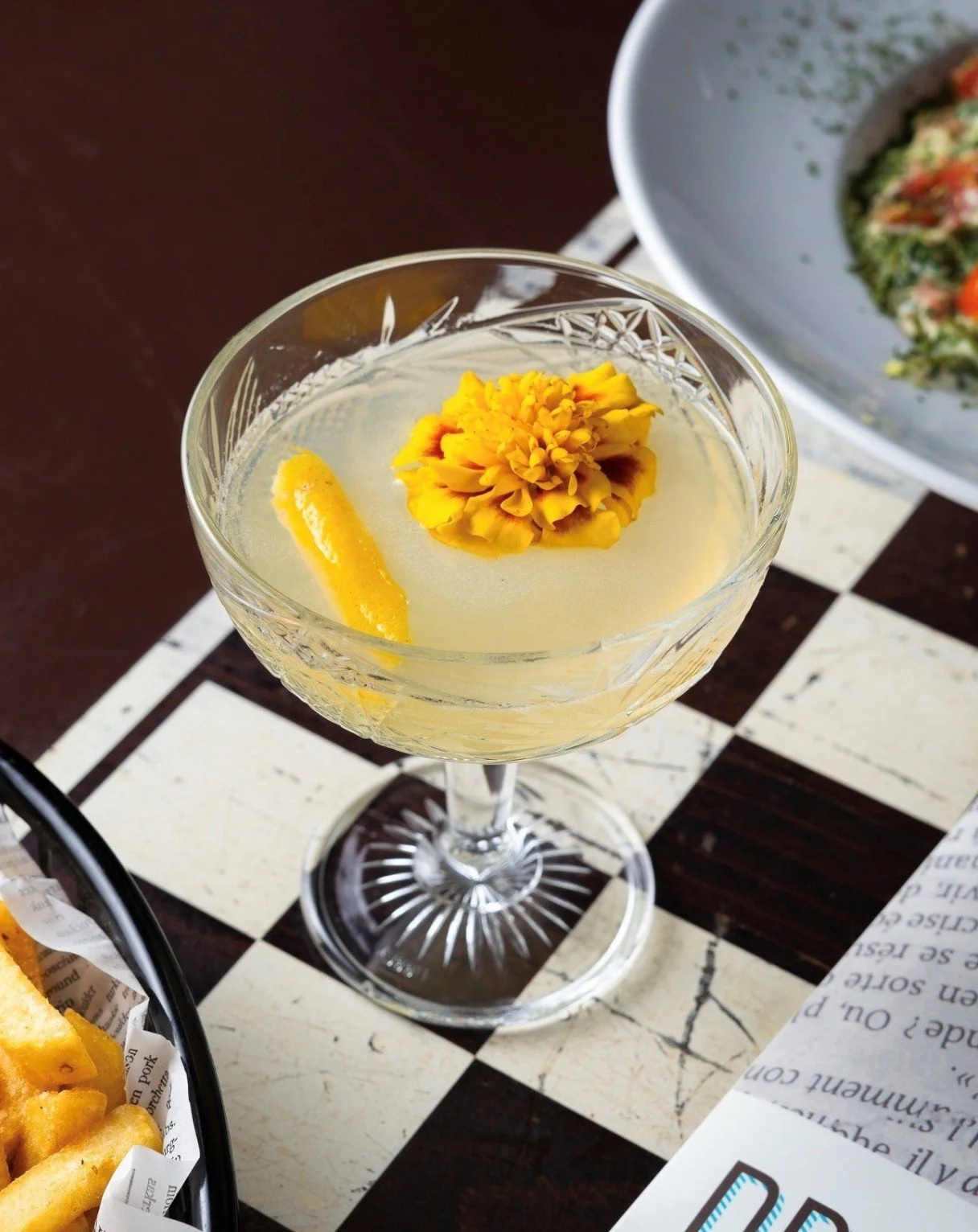 Een cocktail in een glas met een citroenschijfje en een bloemblaadje als garnering, geplaatst op een zwart-wit geruite tafel (schaakbord).