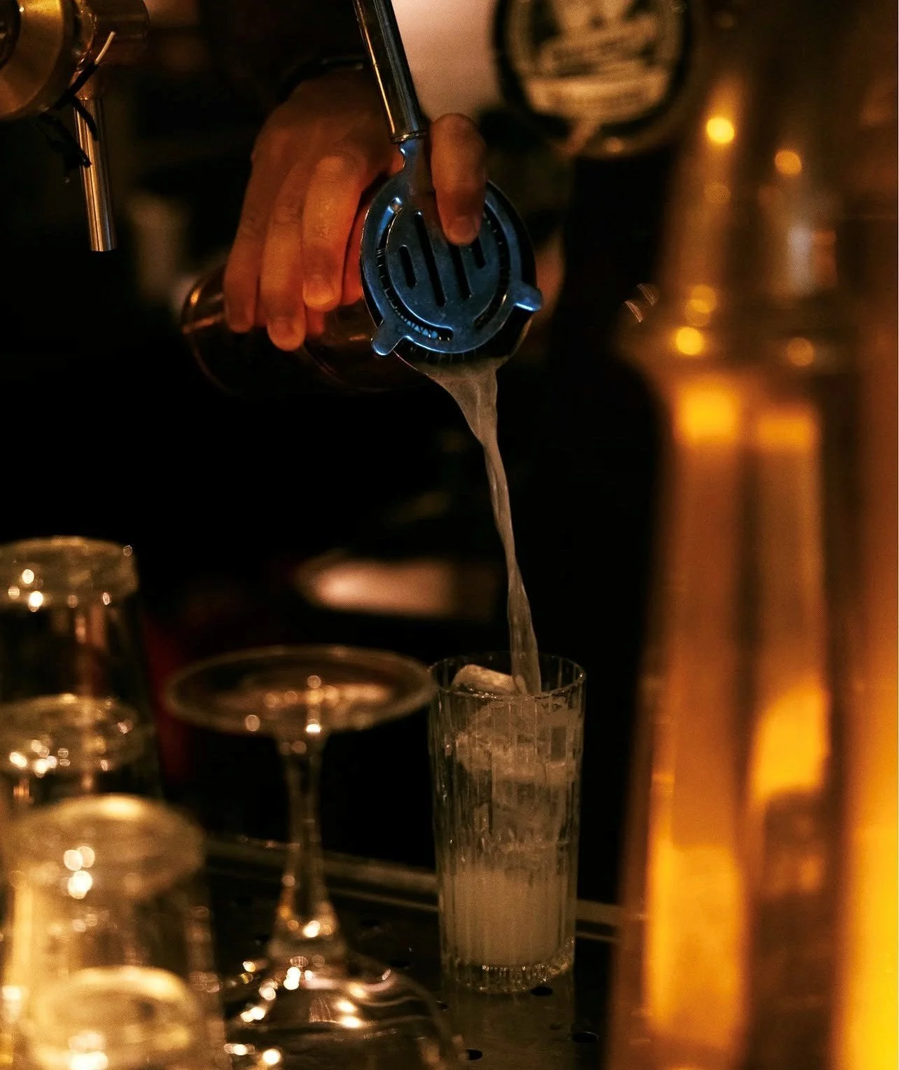 Een hand giet een drankje in een glas in een donkere bar, met meerdere lege glazen op de voorgrond.