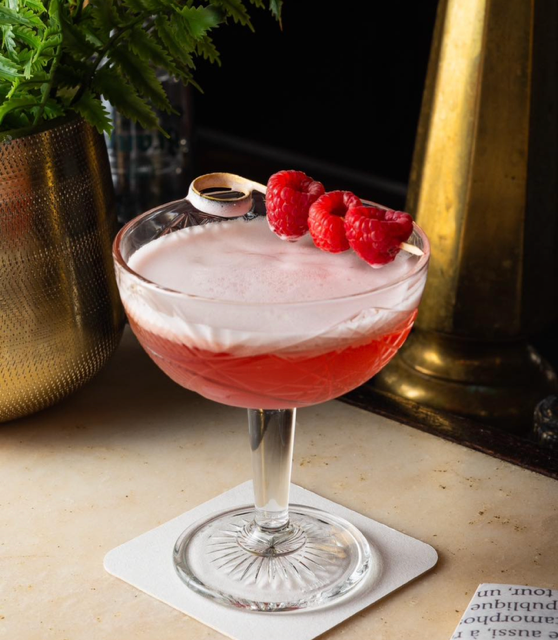 Een roze cocktail met schuimlaag, gegarneerd met frambozen geserveerd in een mooi glas in een warme sfeer.