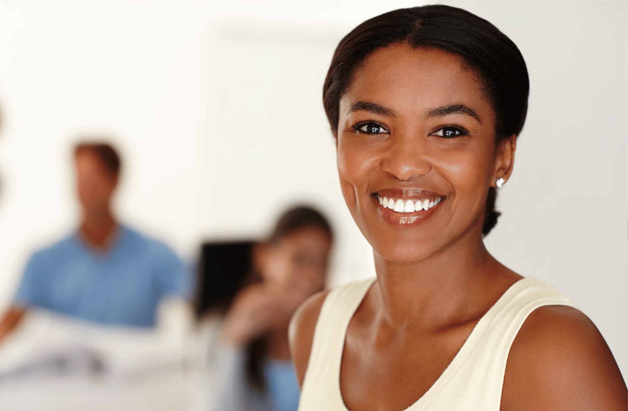 A smiling African American woman with short dark hair, wearing pearl earrings and a light-colored sleeveless top, in a professional setting with blurred colleagues in the background.