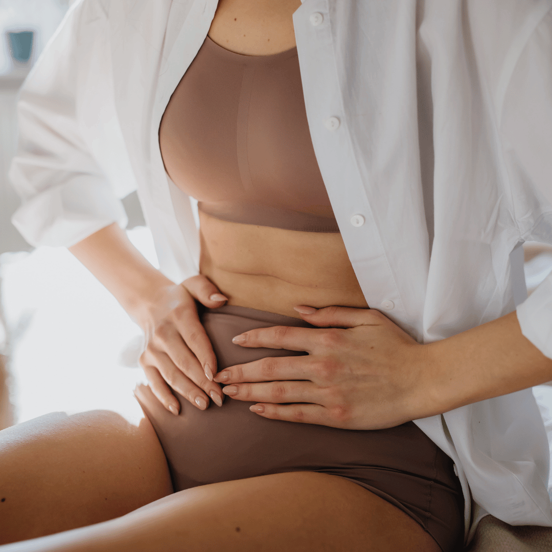 A woman wearing a beige bra and matching underwear, partially covered by an unbuttoned white shirt, sitting indoors with her hand resting on her stomach.