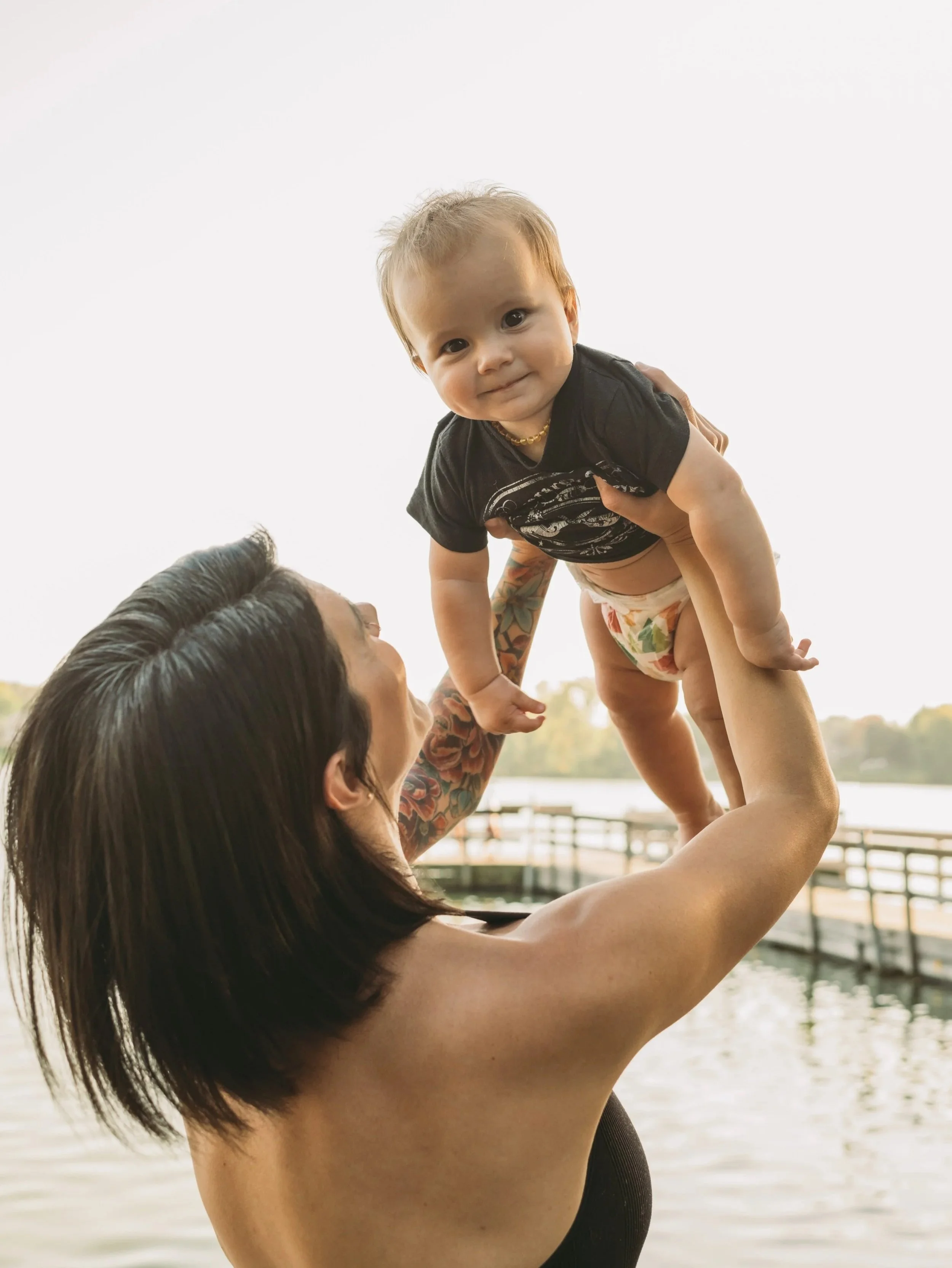 A woman with tattoos on her arm lifting a young boy with light hair and wearing a black shirt and diaper outdoors near water, with trees and a fence in the background.