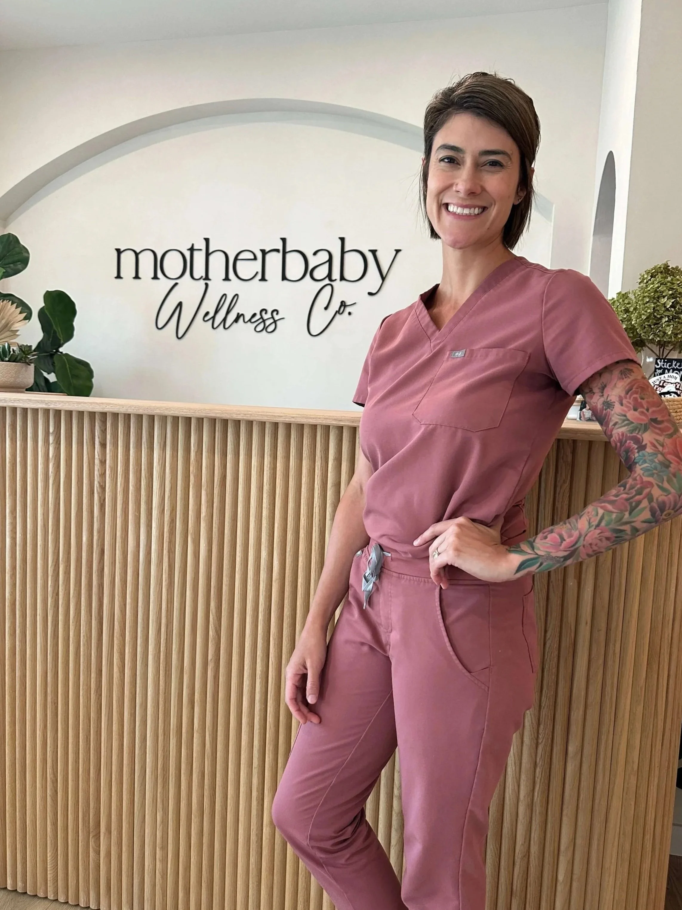 Chrisi Kelly, DC, CACCP, CPM, LM, wearing pink scrubs, standing at a reception desk of MotherBaby Wellness Co.