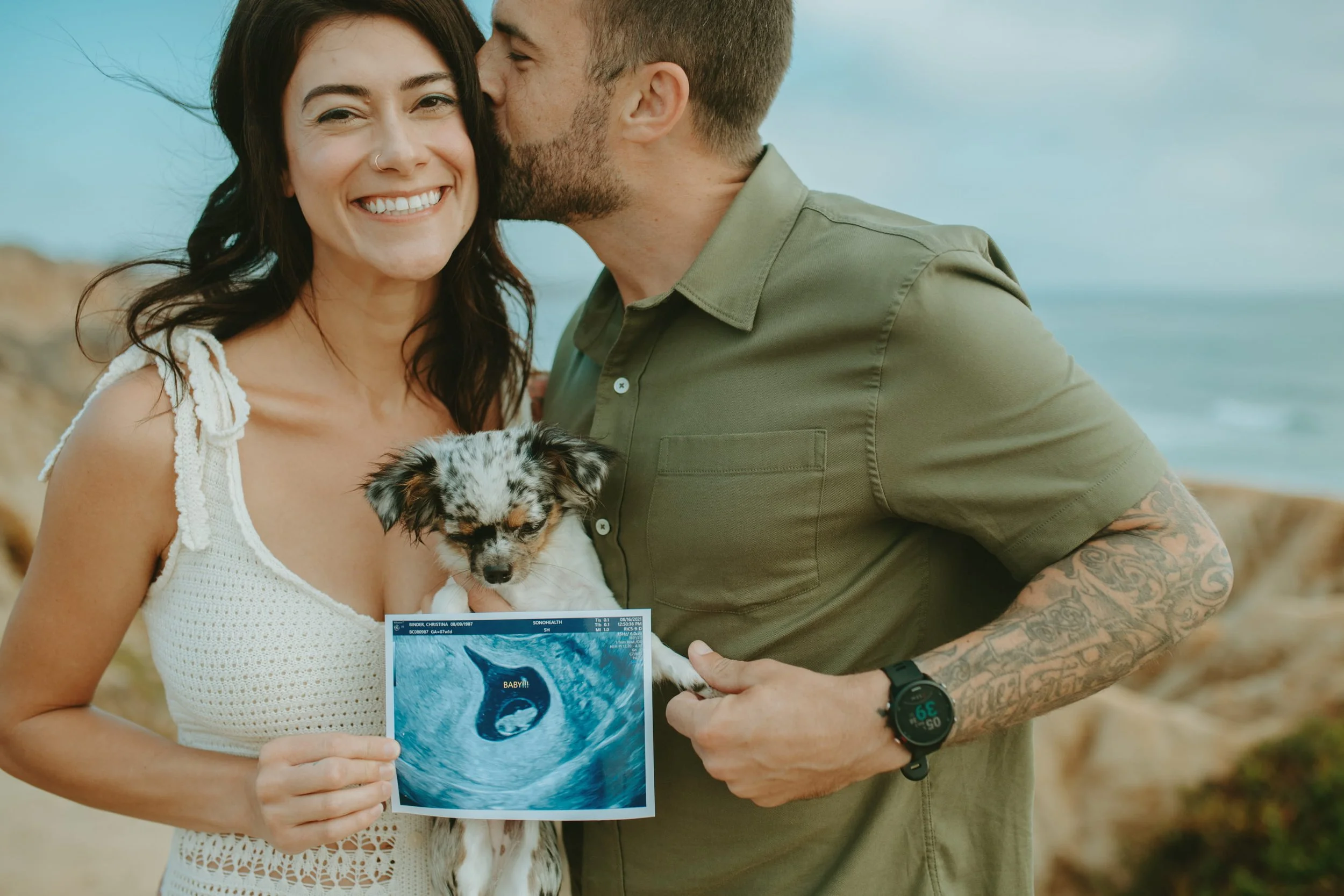 A smiling woman with dark hair holding an ultrasound picture, standing next to a man with tattoos on his arm, who is kissing her on the forehead. The woman is holding a small puppy with a colorful fur pattern, and the background suggests they are at the beach.