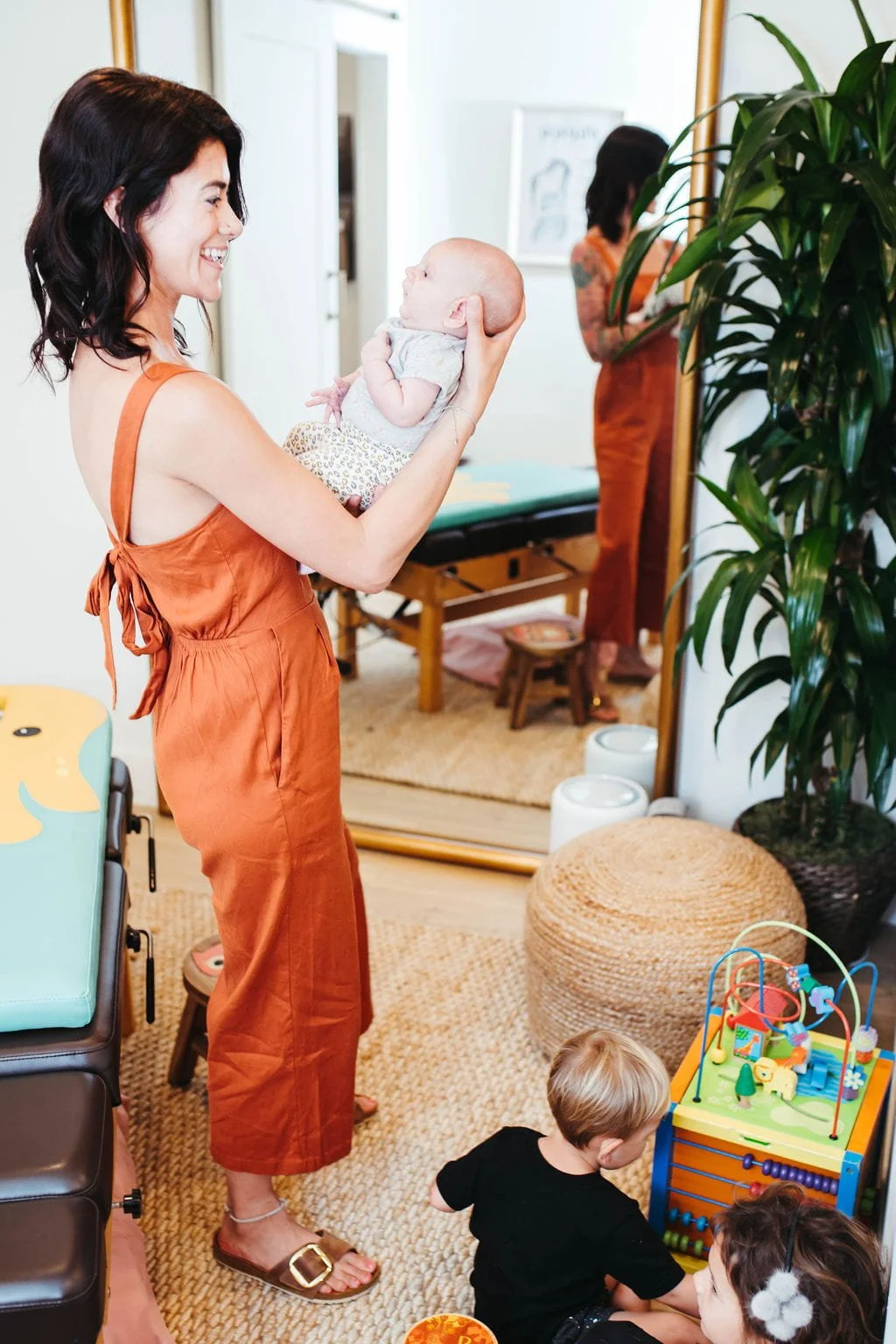 A woman smiling and holding a newborn baby in a room with a mirror, a potted plant, and children playing on the floor.