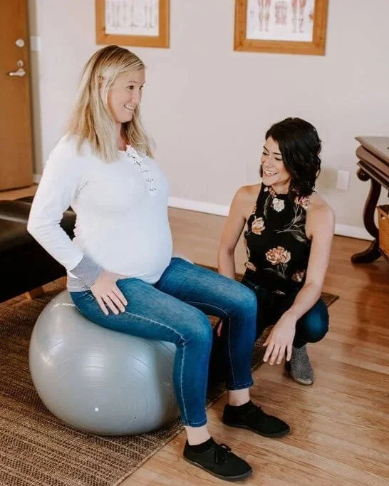 A pregnant woman sitting on an exercise ball, smiling, with a woman kneeling in front of her, inside a room with wooden floors, framed anatomy posters on the wall.