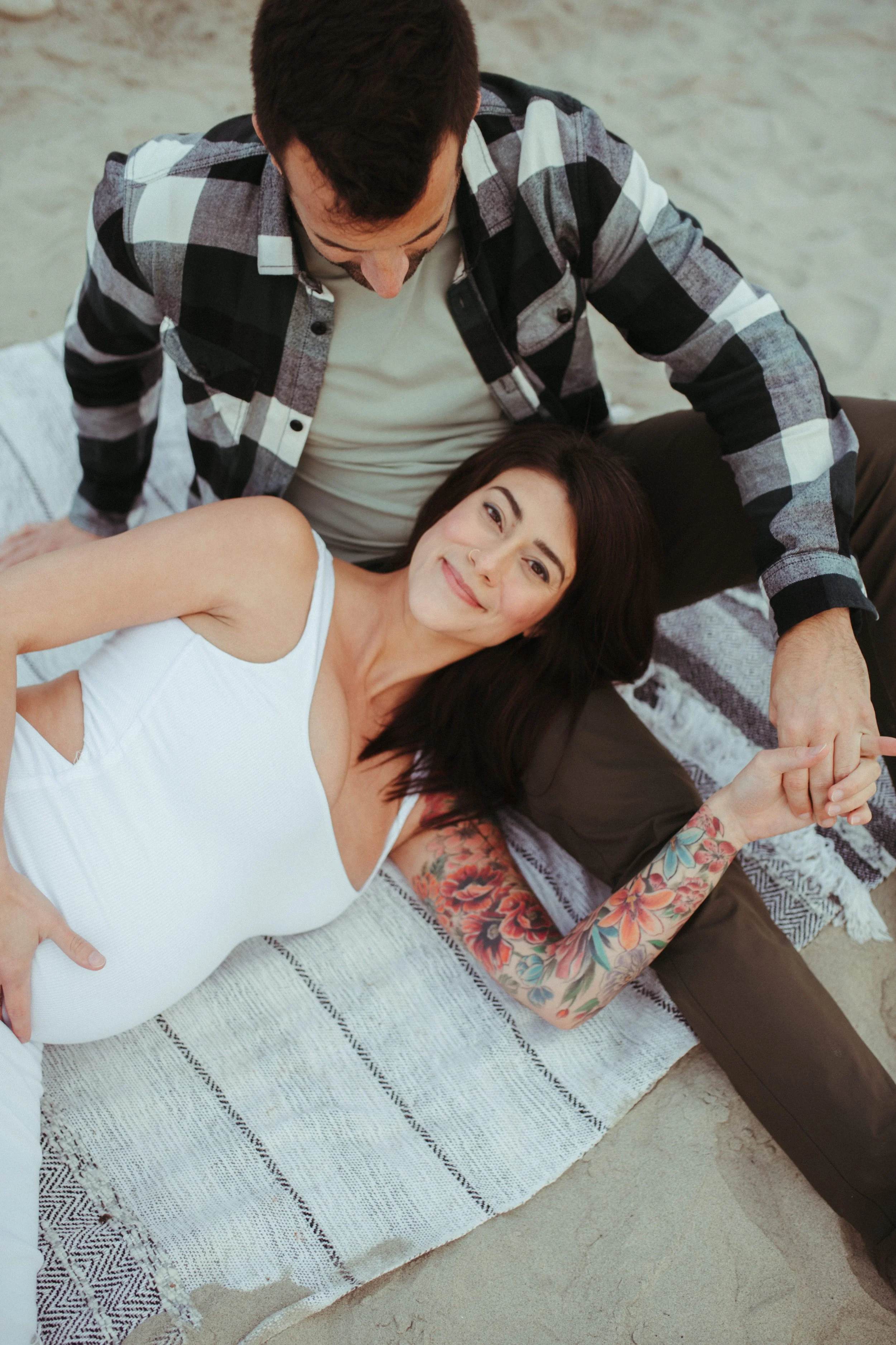 A woman with dark hair and floral tattoos on her arm lying on a blanket on the ground, holding hands with a man with short dark hair, wearing a checkered shirt and a white T-shirt. They are outdoors on a light-colored surface.